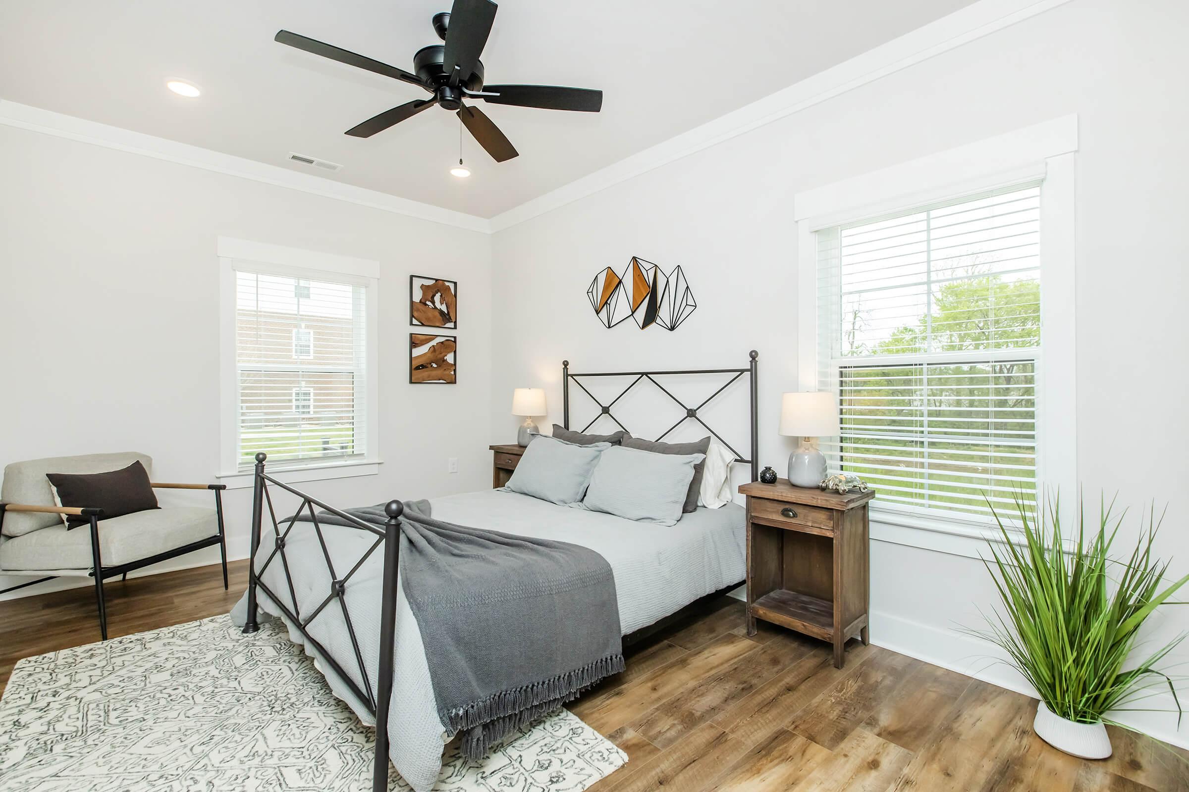 A stylish, modern bedroom featuring a metal bed frame with gray bedding, two bedside tables with lamps, a cozy reading chair, and decorative wall art. Natural light streams in through two windows adorned with white blinds, complemented by a decorative rug and indoor plants, creating a welcoming atmosphere.