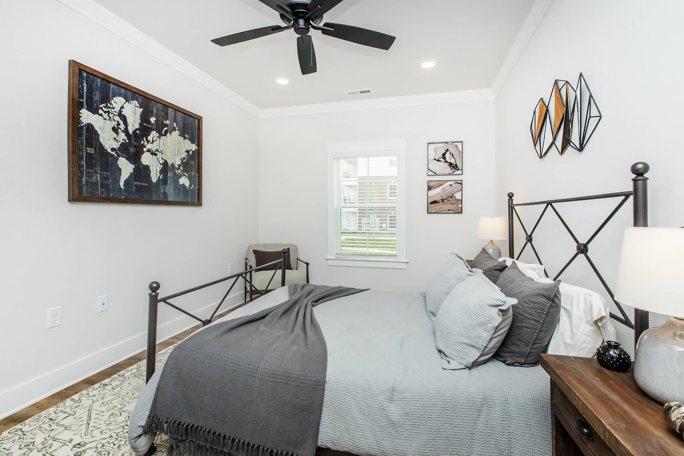 A cozy bedroom featuring a metal bed frame with gray bedding, two bedside lamps, and a wooden nightstand. The wall includes a world map art piece and abstract decorations. A ceiling fan and bright, natural light coming through a window enhance the inviting atmosphere.