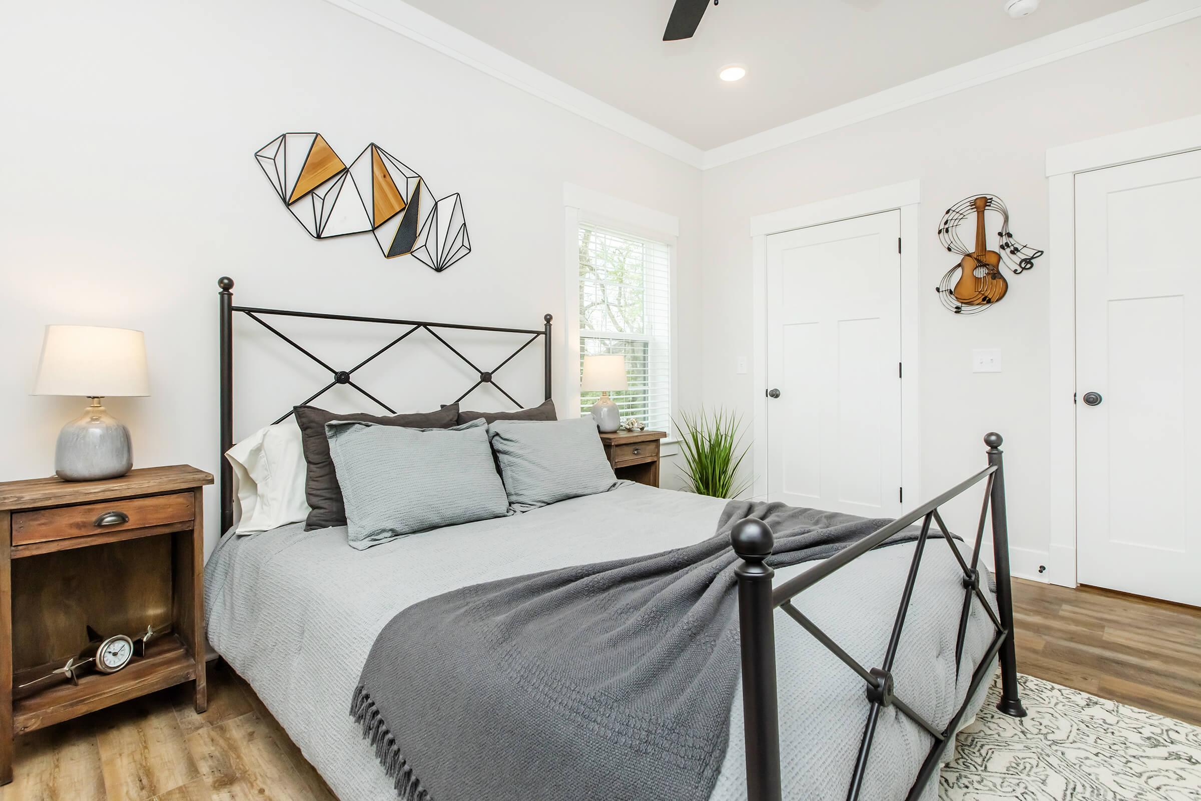 A cozy bedroom featuring a black metal bed frame with gray bedding, two accent pillows, and a textured throw blanket. Beside the bed, there’s a wooden nightstand with a lamp. The walls are adorned with geometric wall art, and a small plant adds a touch of greenery. The room has a calm and inviting atmosphere.