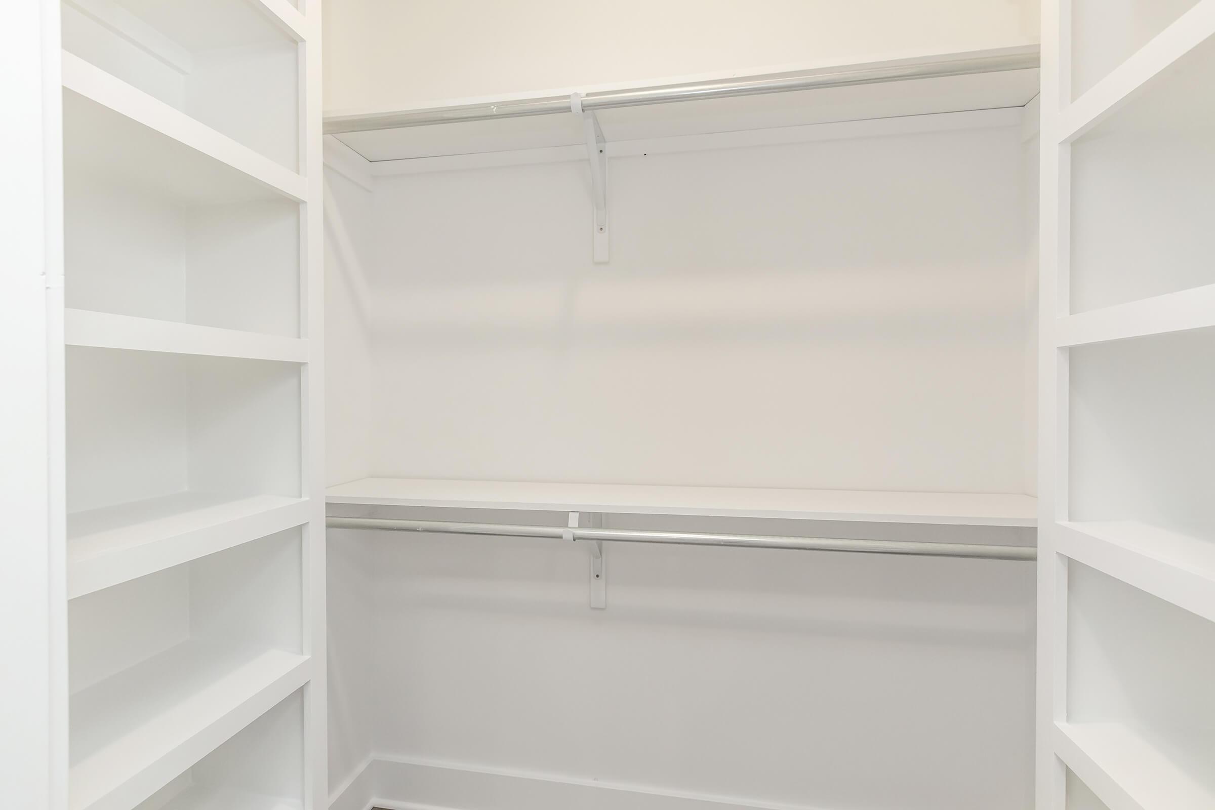 A spacious, empty closet featuring white shelves on both sides and a central hanging rod, all set against a plain white background, showcasing a minimalist design and ample storage space.