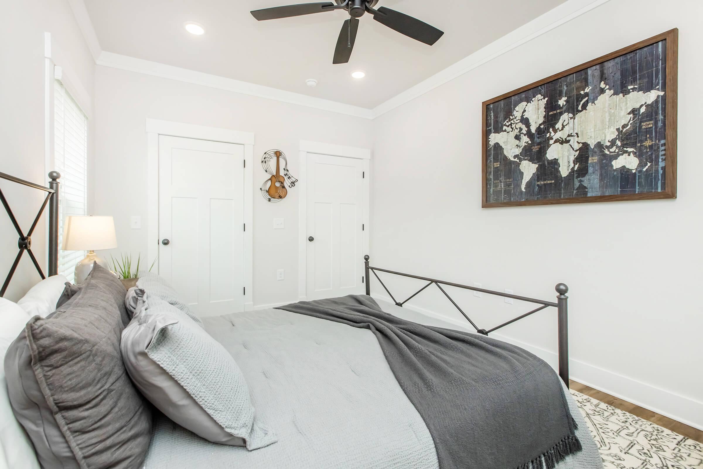 A cozy bedroom featuring a metal bed frame with gray bedding and a gray throw blanket. The walls are painted light, with a large world map artwork hanging above the bed. There are two white doors, a small indoor plant on a nightstand, and a guitar hung on the wall, adding character to the space.
