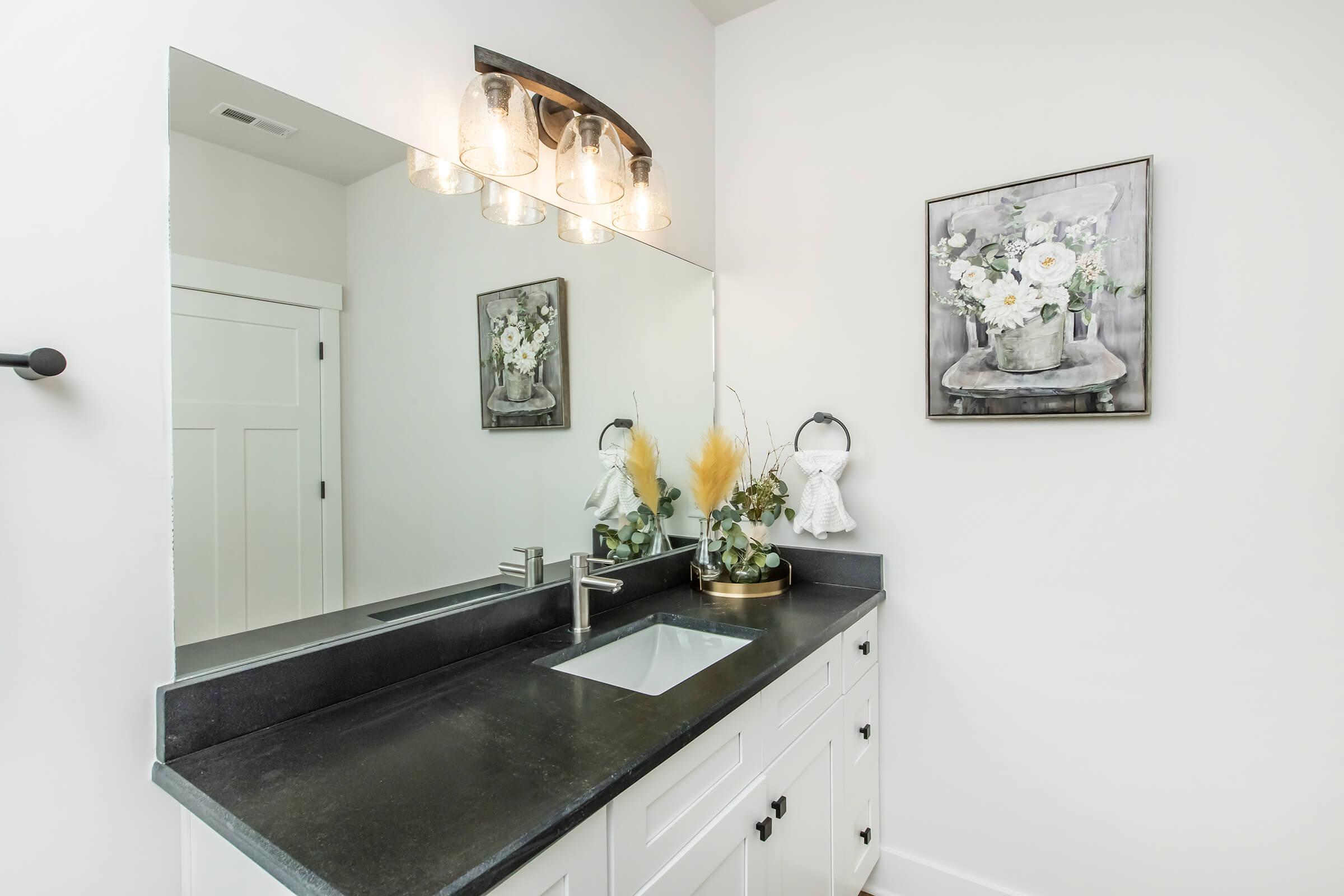 A modern bathroom featuring a sleek dark countertop with a sink, illuminated by stylish pendant lights. A large mirror reflects the space, and there's a framed floral artwork on the wall. Decorative plants and a small vase add a touch of greenery to the minimalist design.