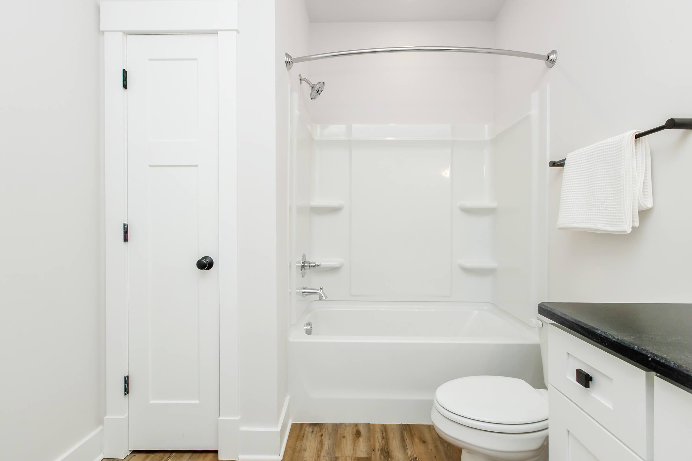A clean, modern bathroom featuring a white bathtub with a curved shower curtain rod, a toilet, and minimalistic storage. Light-colored walls and wood flooring create a bright, airy atmosphere. A towel is neatly hung on a rack beside the bathtub.