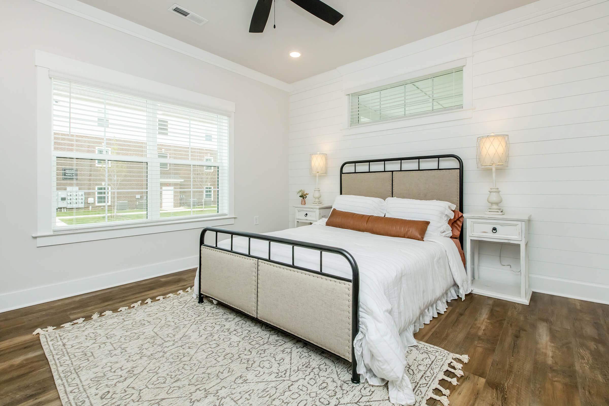 A modern bedroom featuring a large bed with white bedding and a brown accent pillow, two bedside tables with lamps, a decorative area rug, and a window with white blinds allowing natural light to enter. The walls are painted light, and the flooring is hardwood.