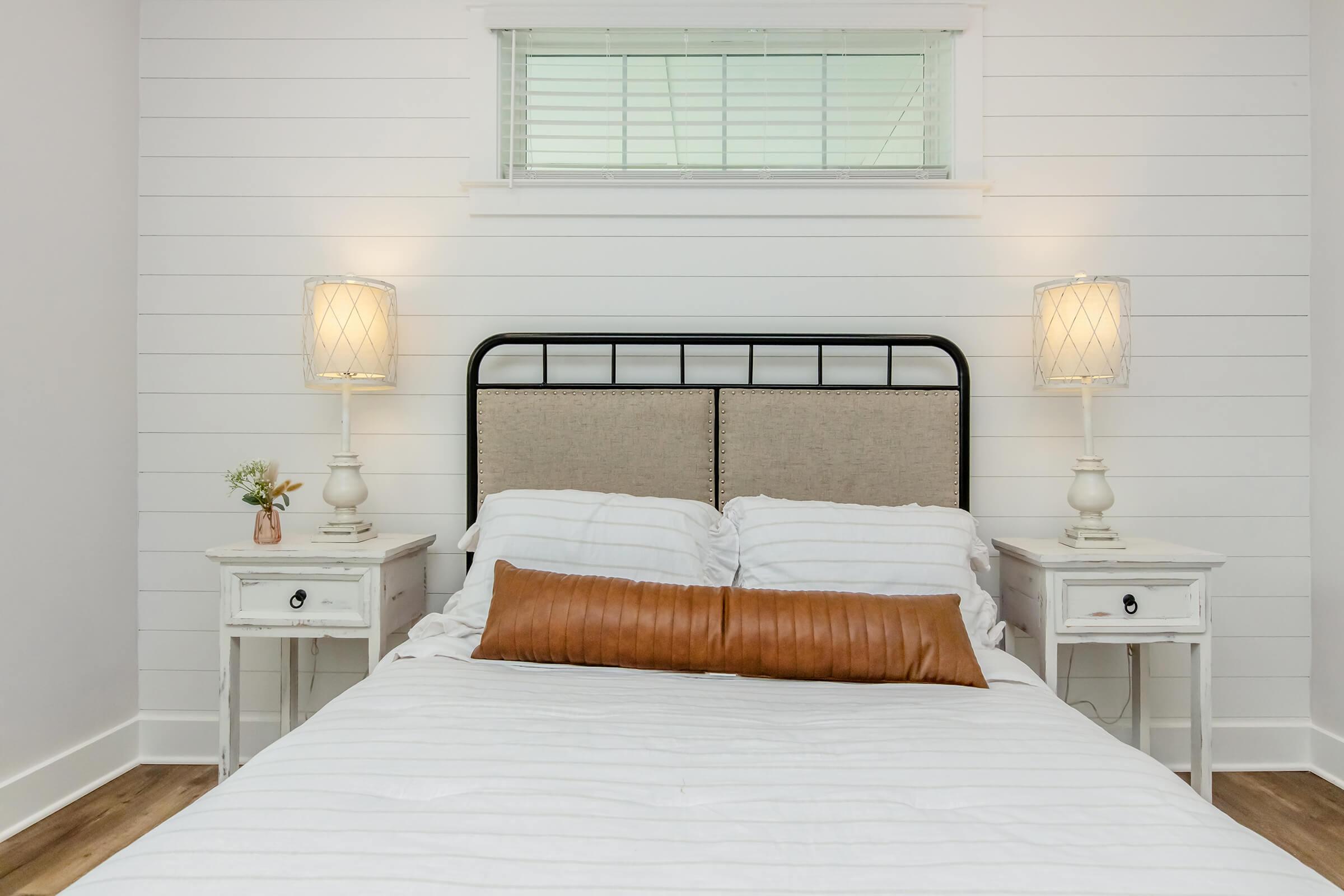 A cozy bedroom featuring a black metal bed frame with a light-colored headboard, white bedding, and a long brown pillow. On either side of the bed are white nightstands with decorative lamps. A small flower vase sits on one nightstand, and a window above provides natural light. The walls are painted white with shiplap accents.
