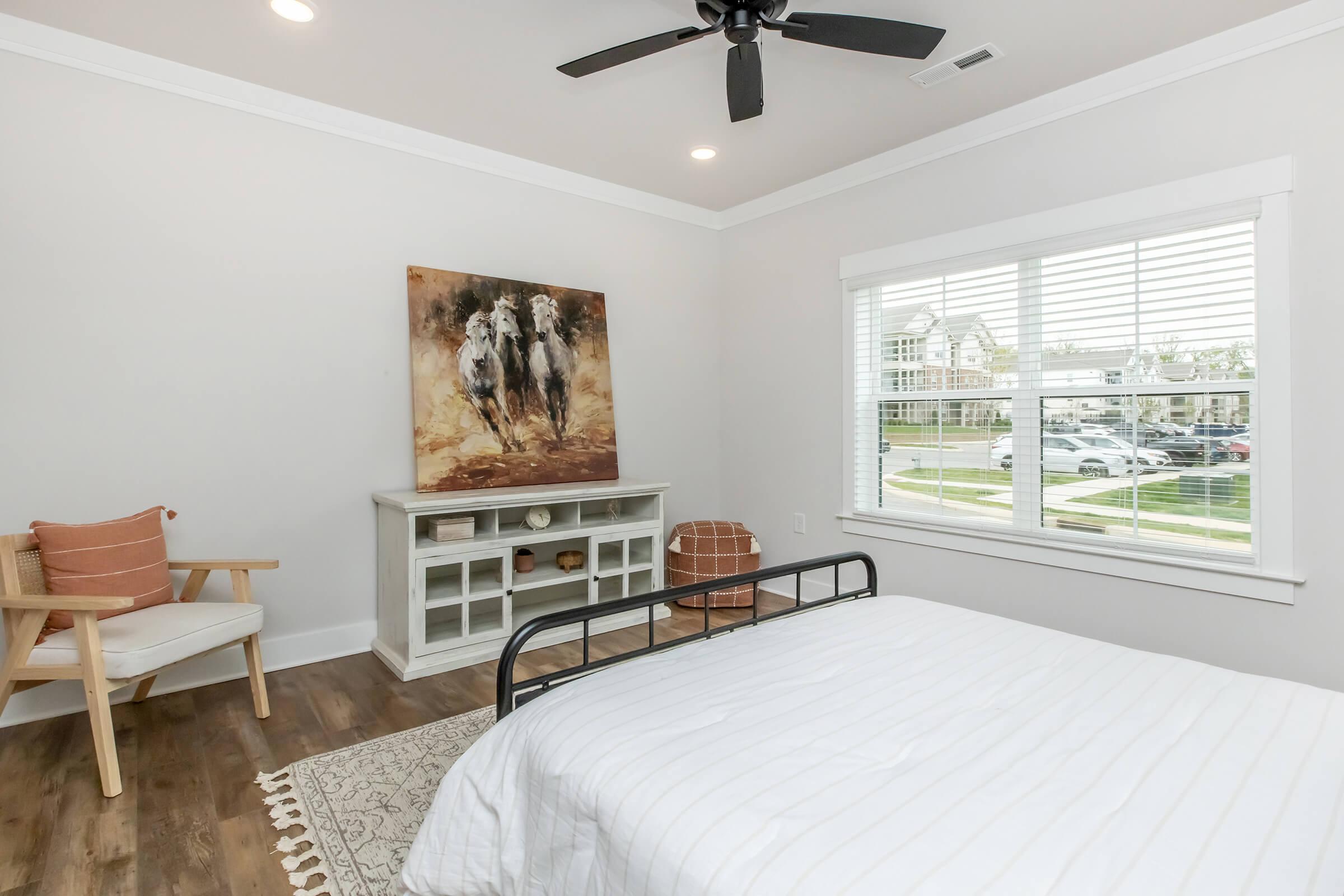 A modern bedroom featuring a black metal bed frame with white bedding, a light wood dresser, and a colorful abstract painting on the wall. The room includes a cozy chair with an orange pillow, a decorative rug, and a window with blinds showing a view of a neighborhood.