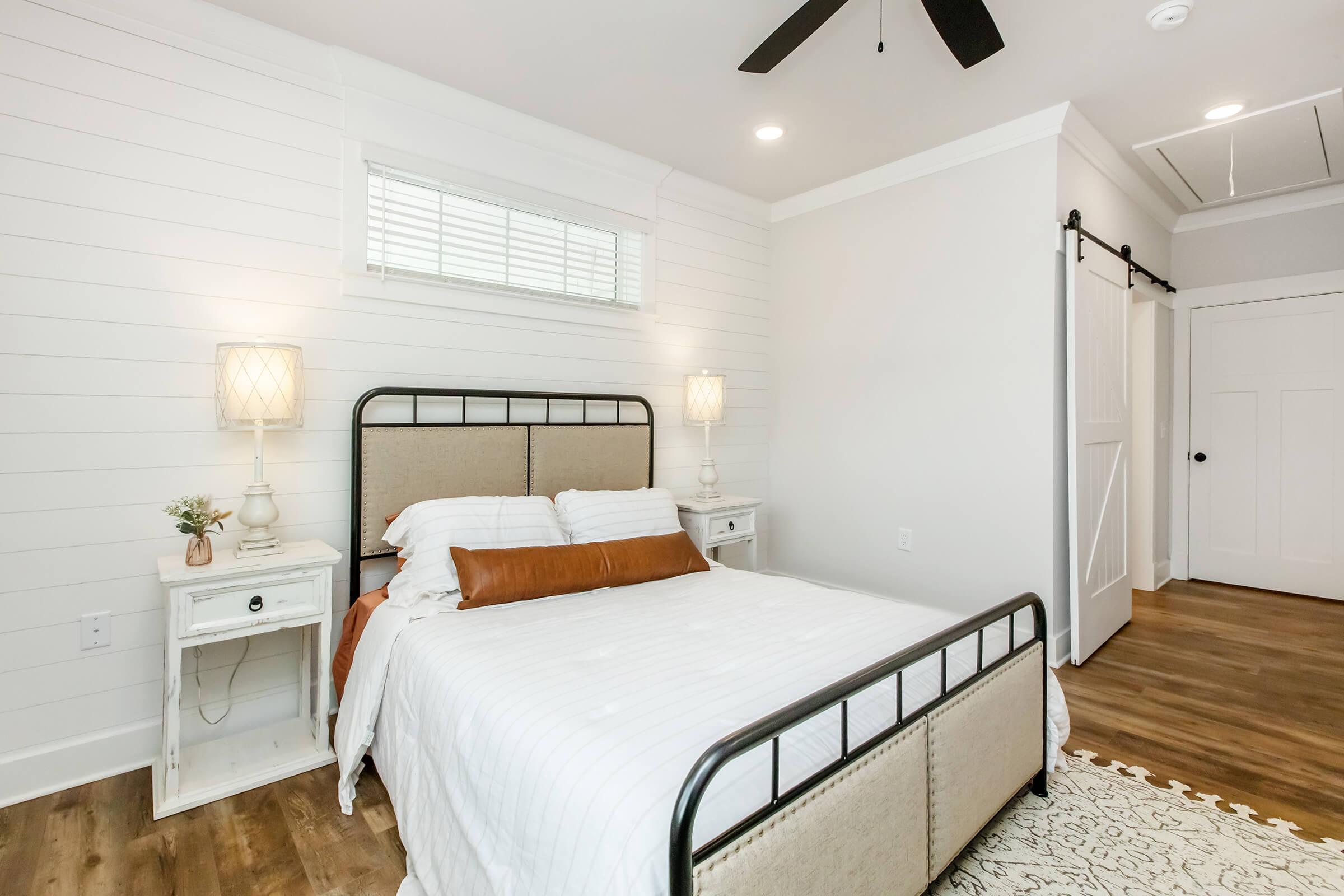 A cozy bedroom featuring a queen-sized bed with a beige and gray headboard, white bedding, and an orange accent pillow. Two nightstands with lamps are on either side of the bed, and there’s a ceiling fan above. The walls are white with wooden paneling, and there is a sliding barn door in the background.