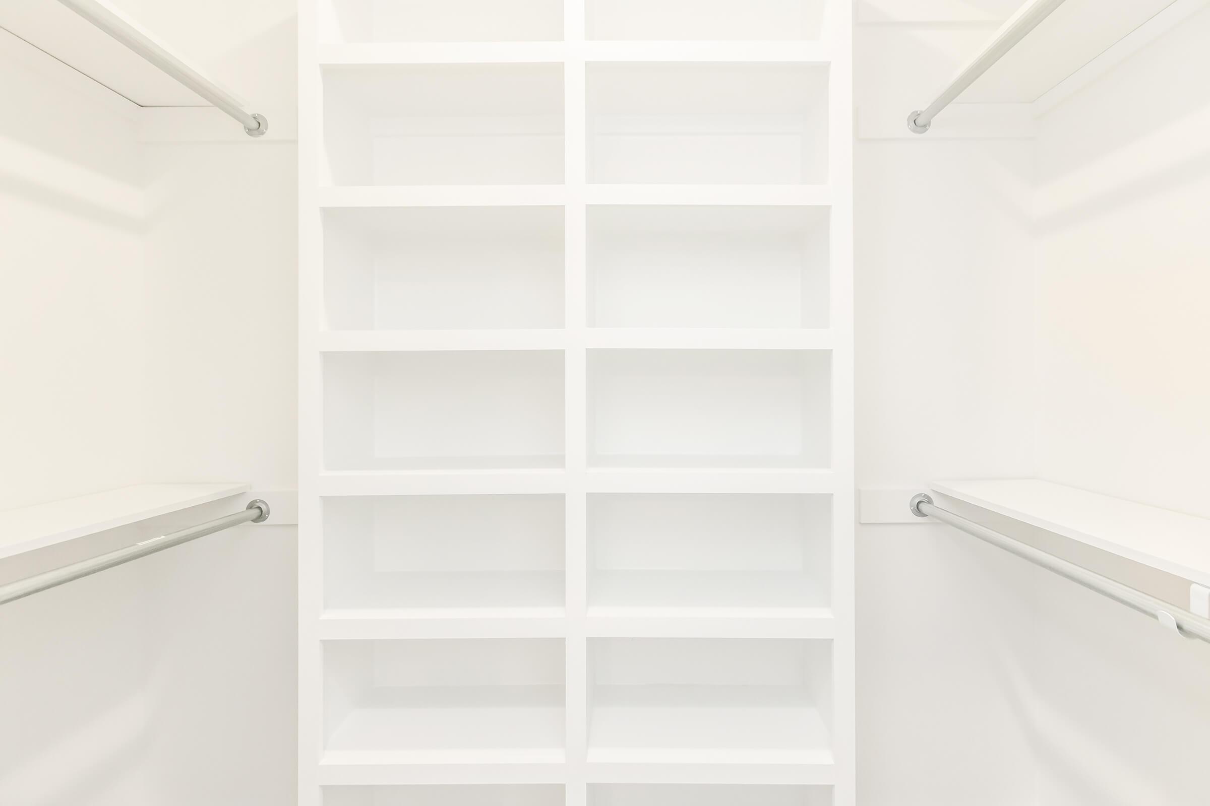 An empty walk-in closet with white shelves and hanging rods, featuring a minimalist design and bright lighting. The shelving is organized into rectangular compartments, providing ample space for storage. The walls and shelving are clean and bright, creating a spacious and airy feel.