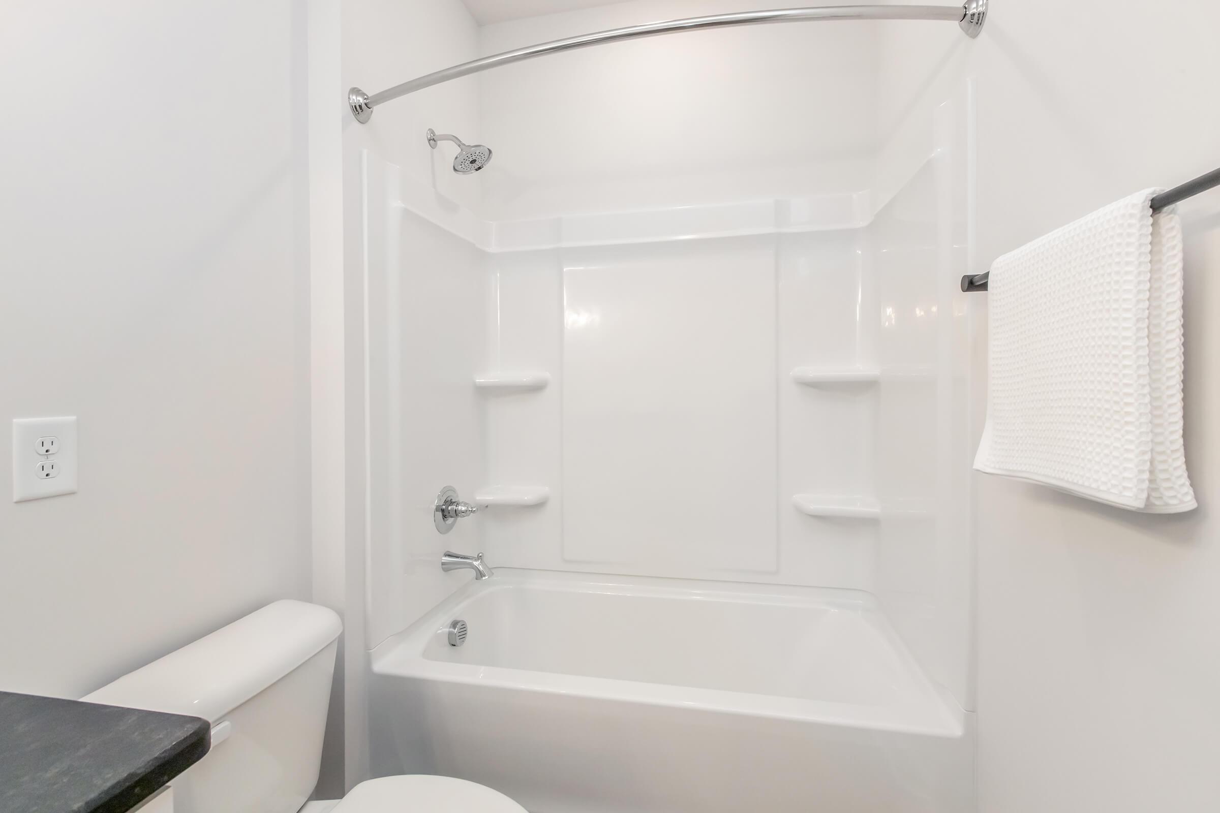 A clean and modern bathroom featuring a white bathtub with a shower, a chrome faucet, built-in shelves for storage, and a white towel hanging on a rack. The walls are painted in a light color, and there is a white toilet visible in the corner.