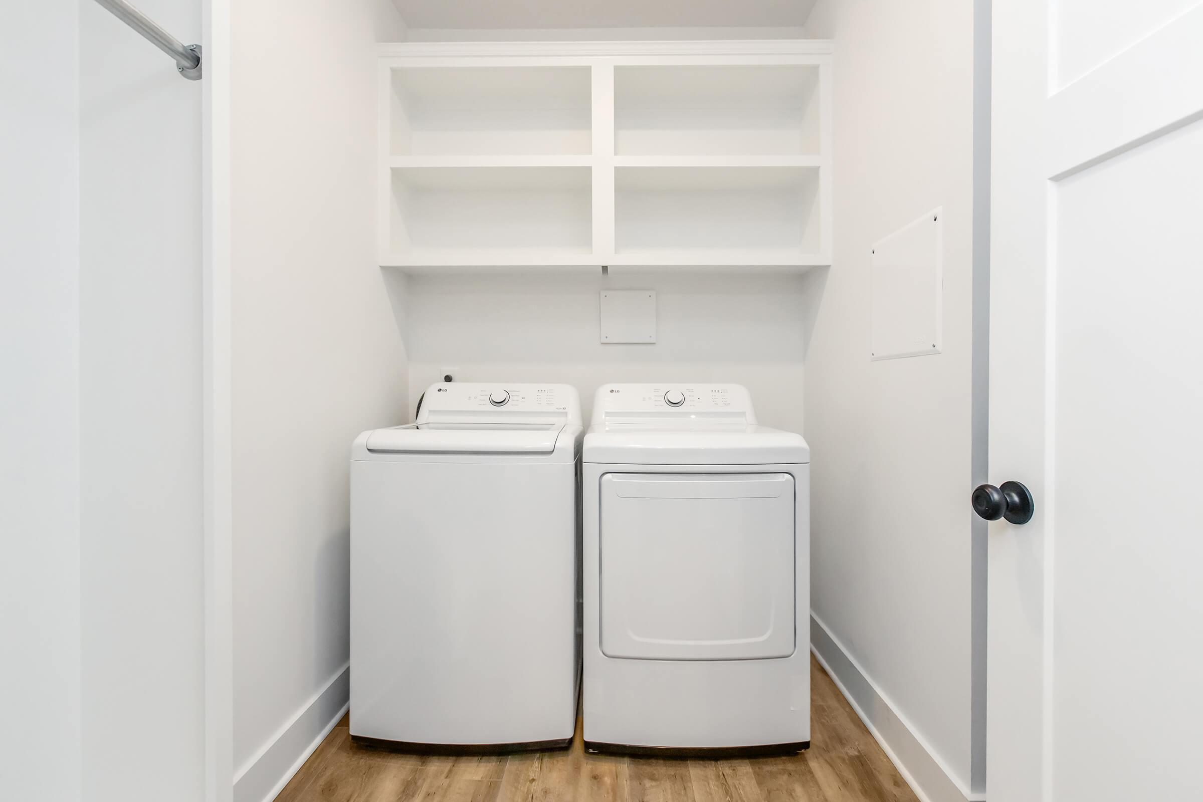 A clean laundry room featuring a white washing machine and dryer side by side. Above them, there are white shelves for storage, and the walls are painted in a light color. The floor is wood-like, adding warmth to the space. A door is visible on the right, leading out of the room.