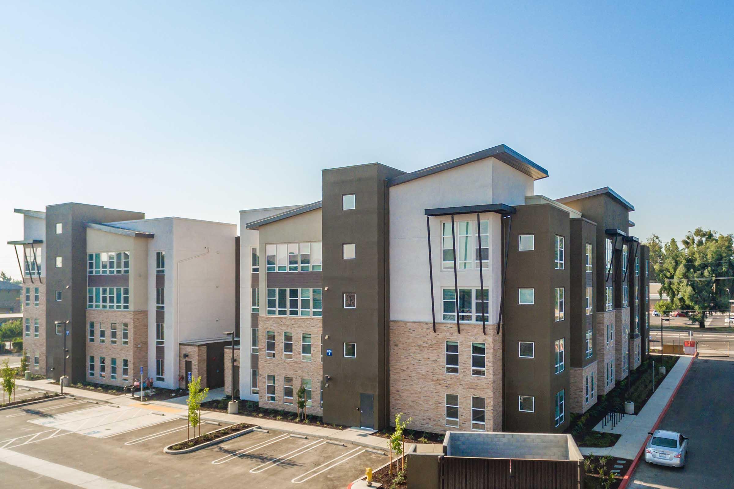 A modern multi-story apartment building with a mix of brick and stucco exterior. The structure features large windows and a contemporary design. An empty parking lot is in the foreground, and clear blue skies are visible above.