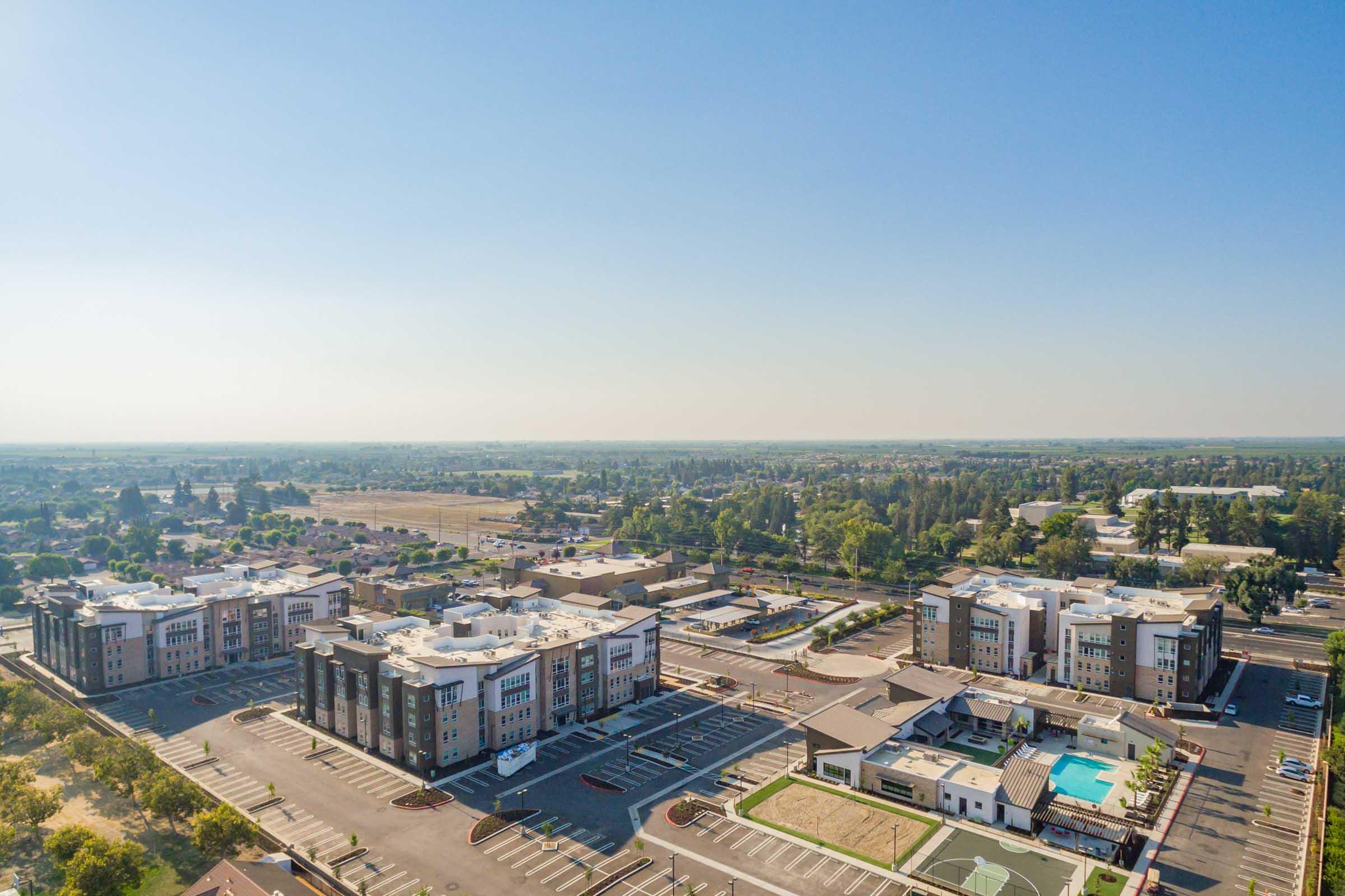 Aerial view of a modern multi-building complex with three large structures, parking lots, and a pool area. Surrounding landscape features greenery and open spaces, under a clear blue sky. The area appears to be part of a suburban setting with visible roads and neighborhoods in the distance.