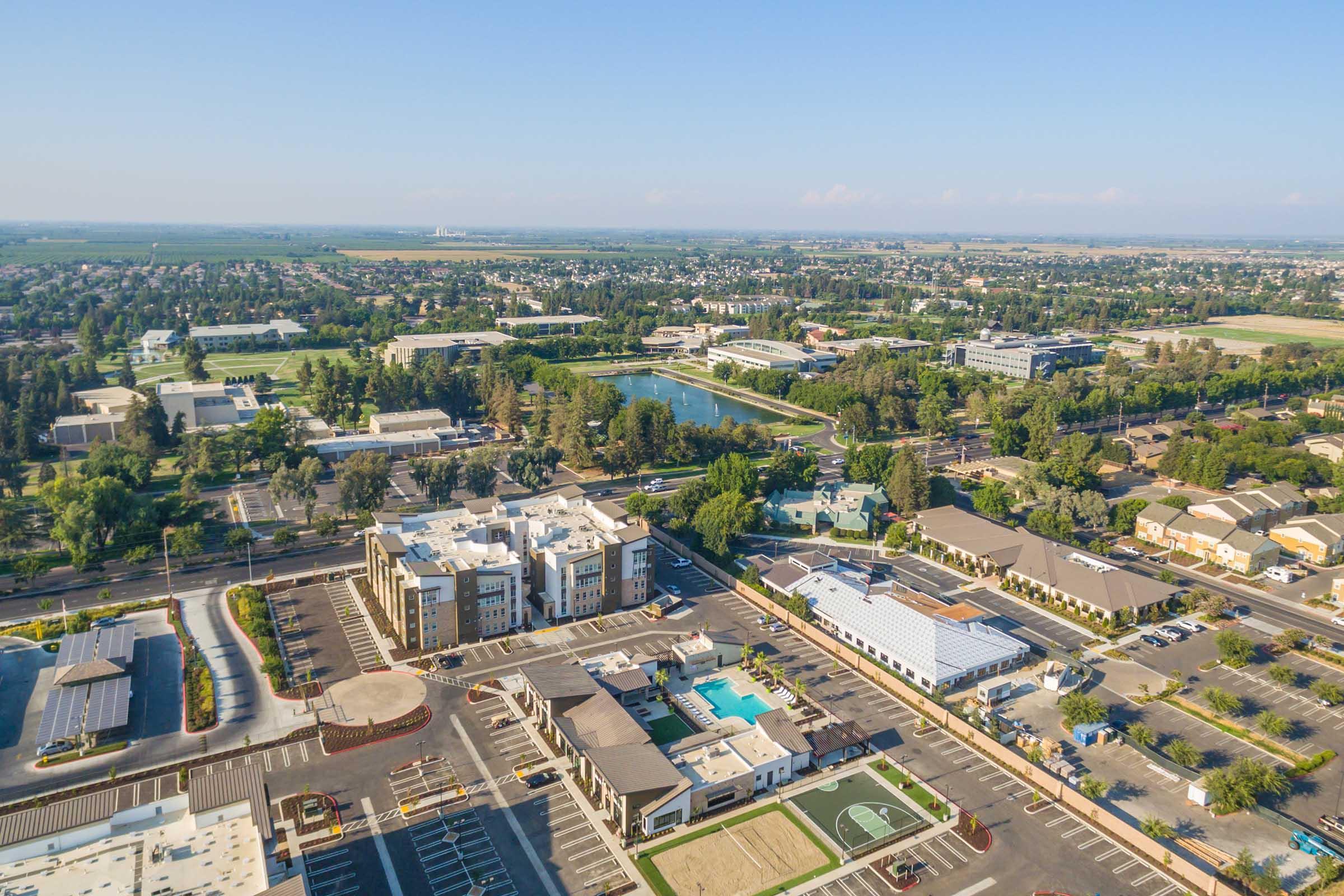 Aerial view of a suburban area featuring residential buildings, a swimming pool, and sports courts. In the background, green fields and trees are visible, along with other structures and a small body of water. The landscape is clear and expansive, indicating a developed community with plenty of open space.