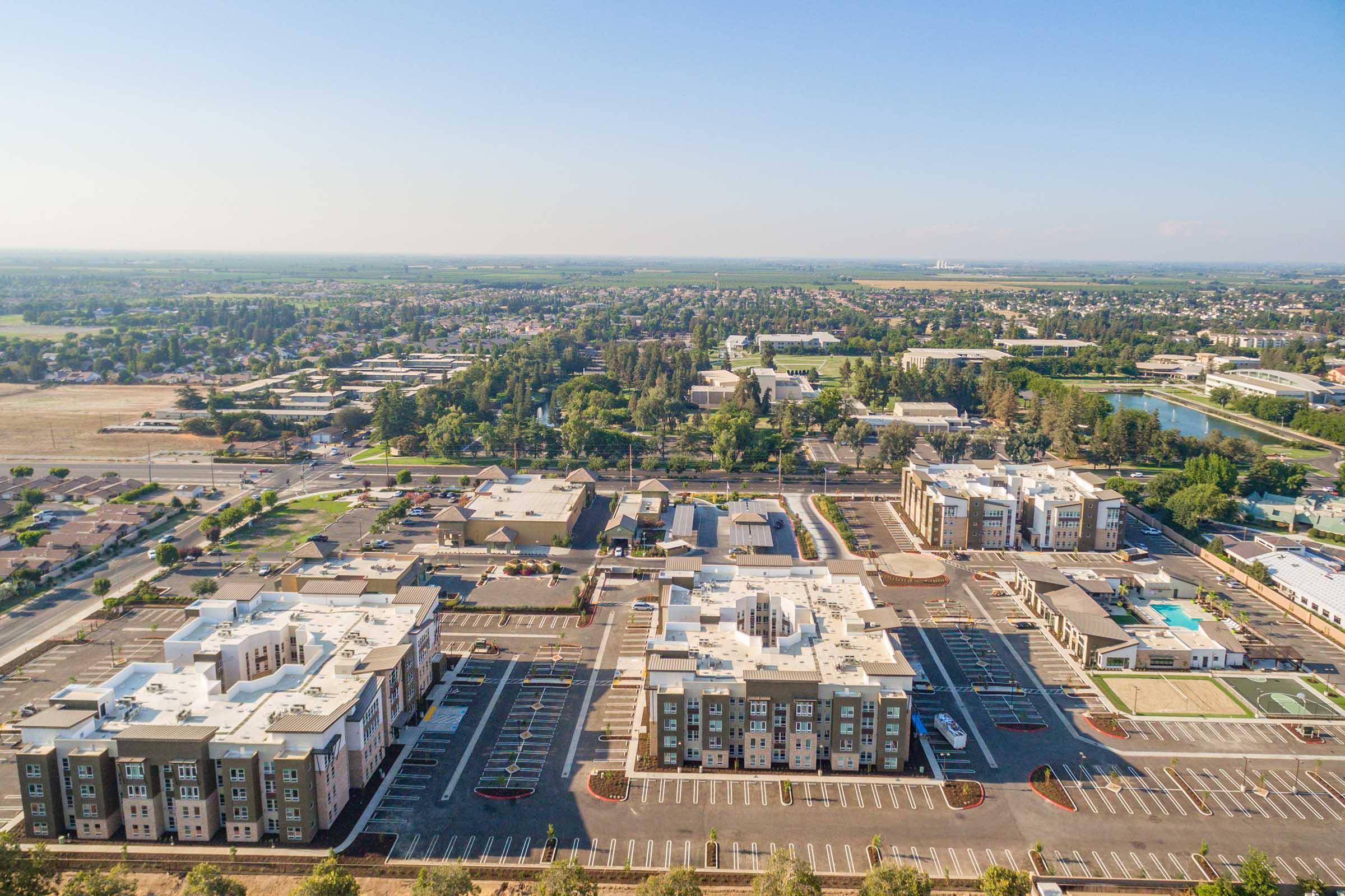 Aerial view of a suburban area featuring modern apartment buildings and an expansive parking lot. The landscape includes low-rise buildings, roads, and greenery, with a residential and commercial mix. In the background, rolling hills and further development are visible under a clear blue sky.