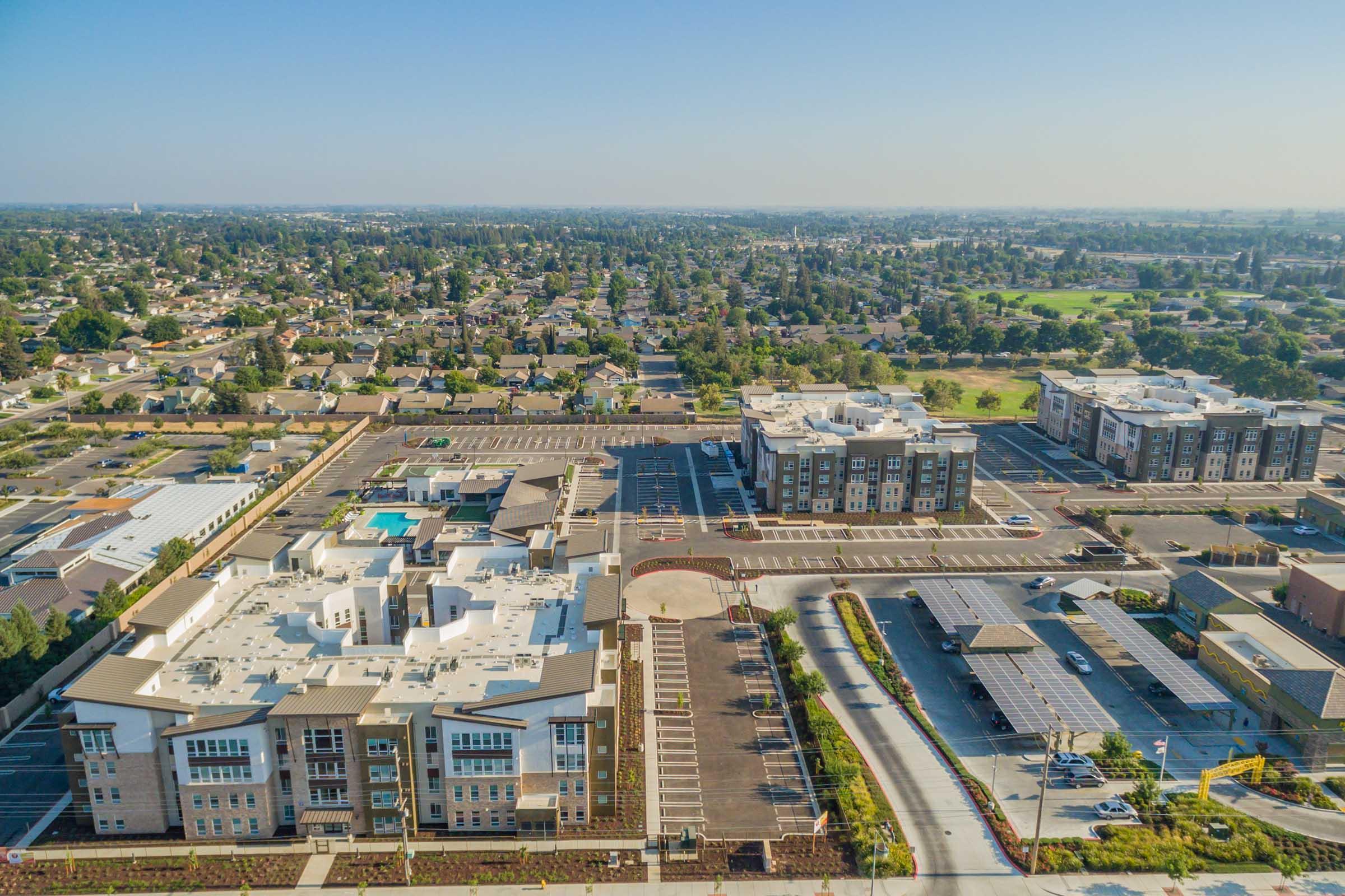 Aerial view of a suburban area featuring several modern apartment buildings, a large parking lot, and a swimming pool. The landscape includes lush green spaces and tree-lined streets, with additional residential and commercial buildings visible in the background.