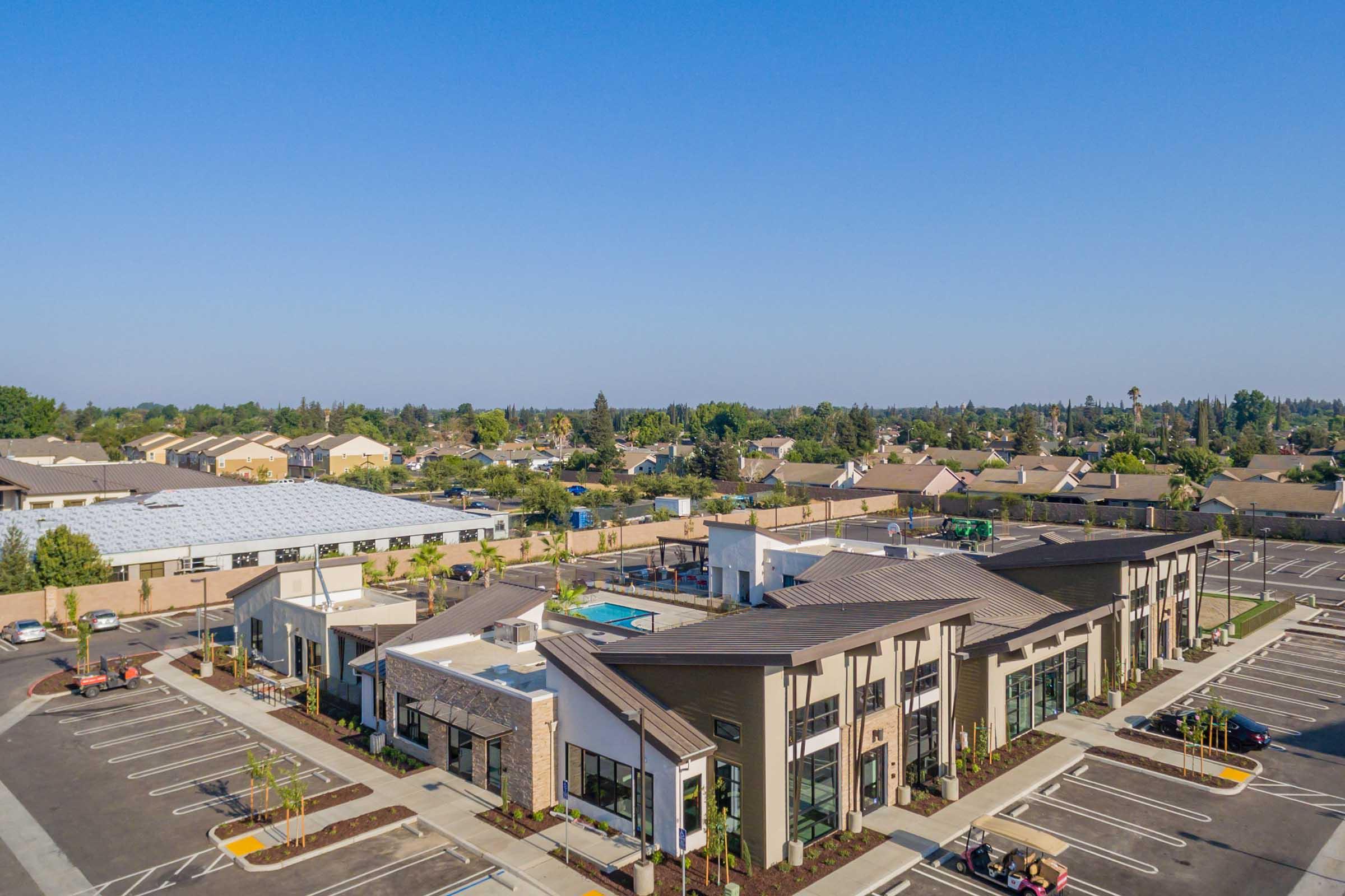 Aerial view of a commercial building complex with modern architecture, featuring a swimming pool, ample parking space, and greenery. Surrounding the complex are residential homes and a clear blue sky, indicating a sunny day.
