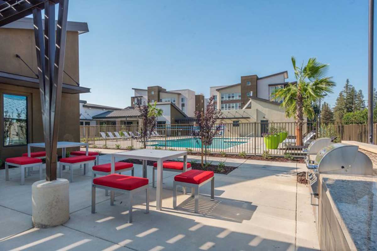 A sunny outdoor area featuring a pool surrounded by lounge chairs, with a patio that includes modern tables and red seating. Palm trees and a barbecue grill enhance the relaxing atmosphere, creating an inviting space for social gatherings.