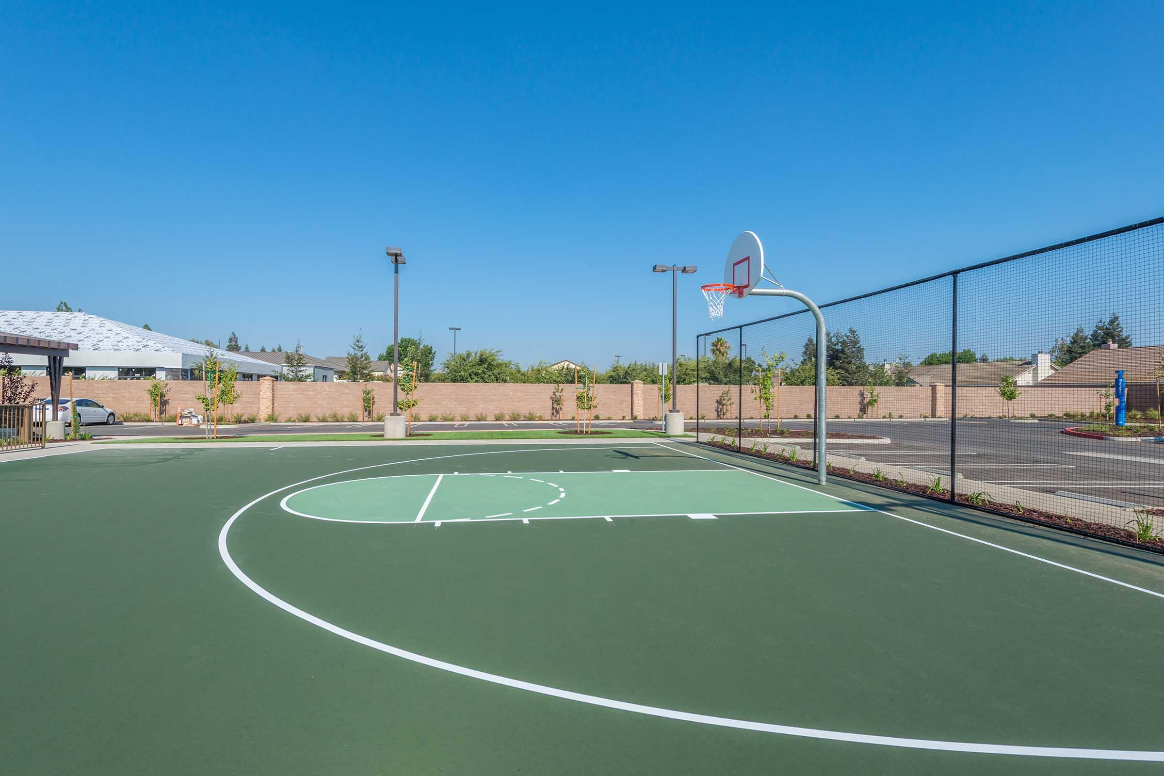 A basketball court featuring a green surface, marked with court lines, a basketball hoop at one end, and a backdrop of a clear blue sky. Surrounding the court are short walls and landscaped areas, creating a spacious outdoor setting ideal for playing basketball.