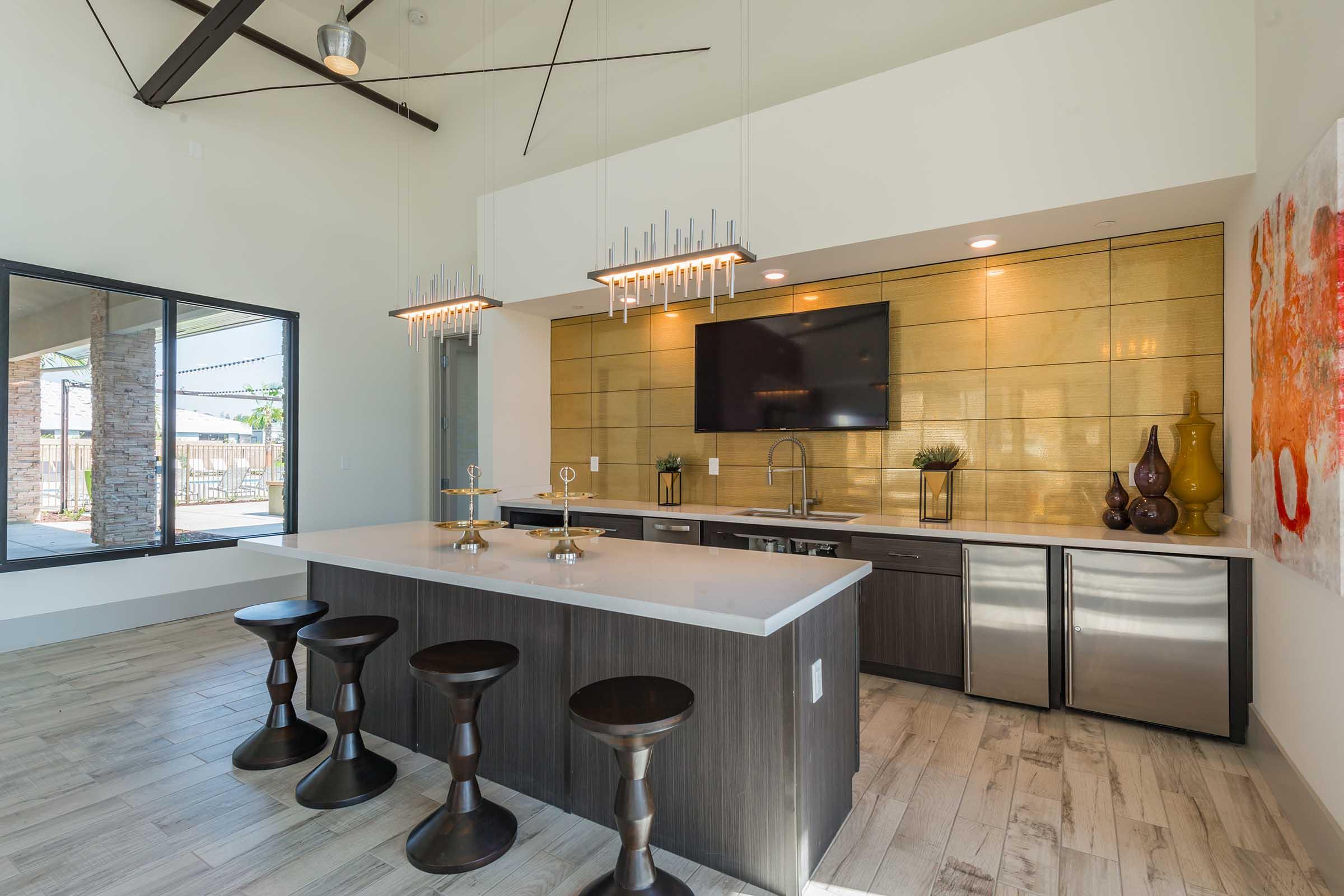 Modern kitchen with a large central island featuring dark wood stools. The area has gold-tone backsplash, stainless steel appliances, and decorative vases. Brightly lit with two stylish pendant lights and a large window providing natural light, showcasing an outdoor view. Contemporary art decorates the wall.