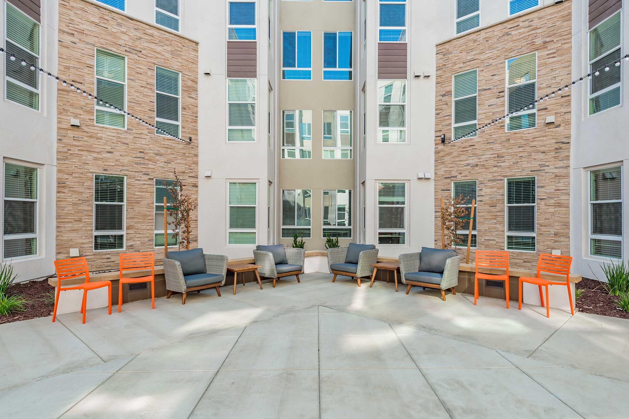 A modern indoor courtyard with stylish seating arrangements. The space features gray sofas, orange chairs, and decorative plants, surrounded by building walls with multiple windows. The overall design is bright and inviting, ideal for socializing or relaxation.