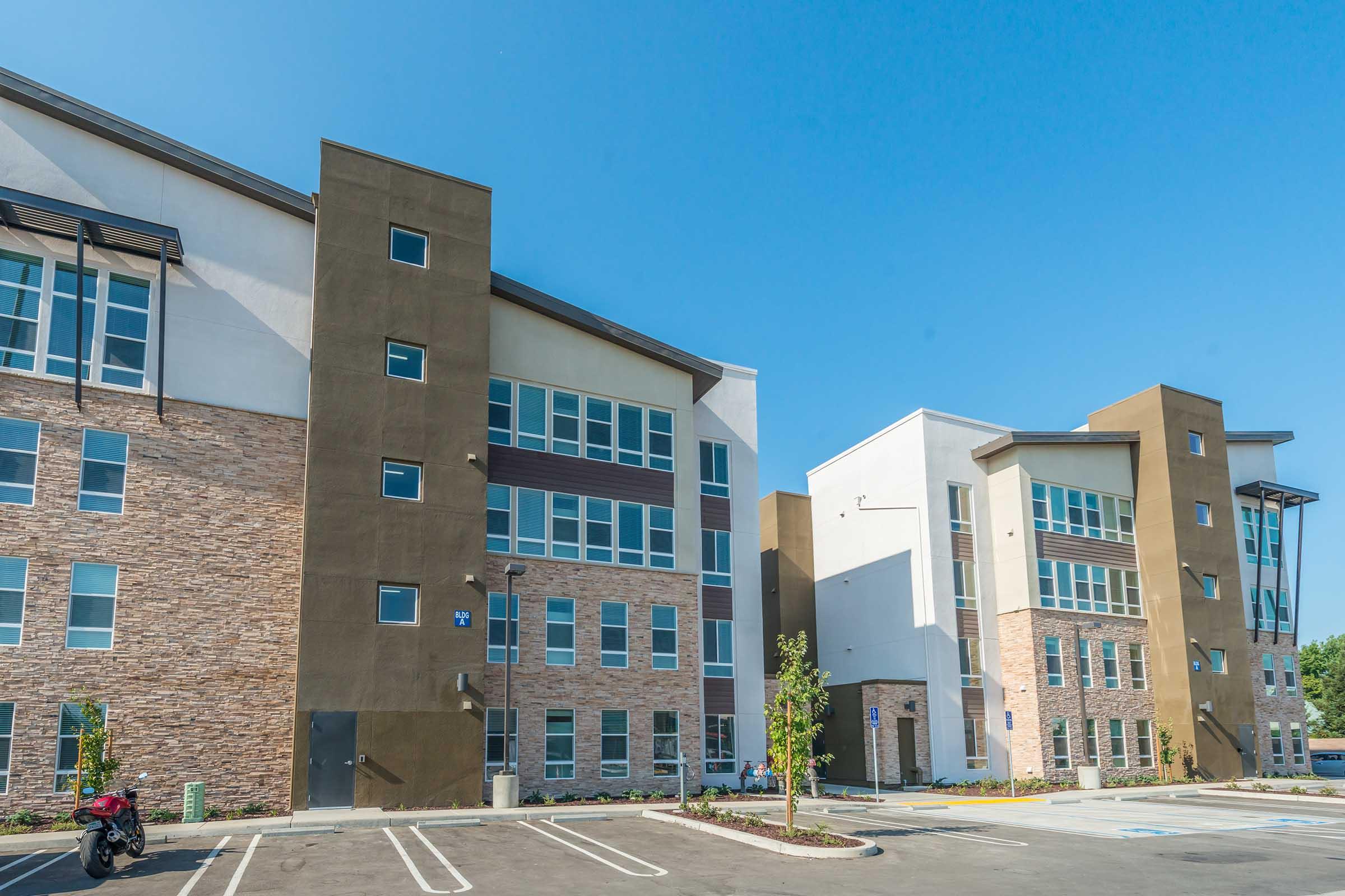 Two modern multi-story residential buildings with a mix of beige stone and brown panels. The structures have large windows and are set against a clear blue sky. An empty parking lot is visible in front, featuring a few small trees and shrubs. The overall atmosphere is bright and inviting.