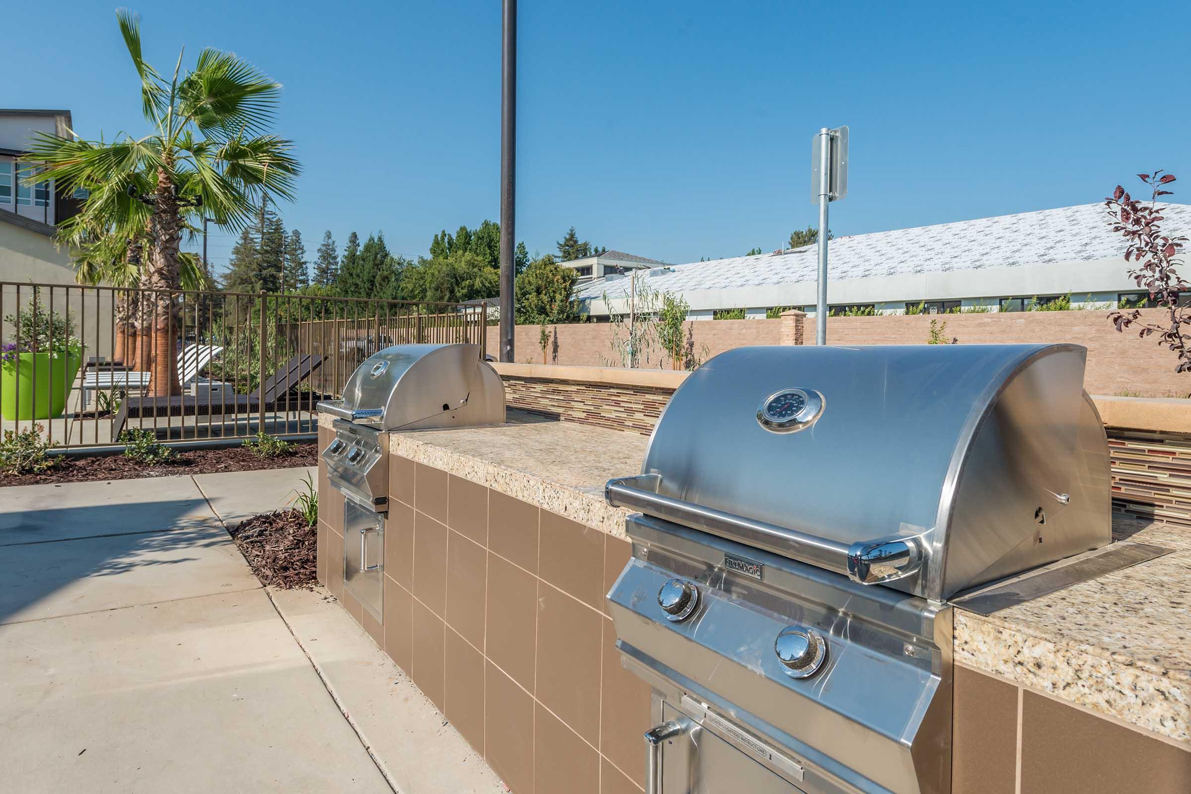 Two stainless steel outdoor grills set on a stone countertop in a sunny patio area. A palm tree is visible nearby, and a modern building can be seen in the background. The space is well-maintained, with landscaping and a clear blue sky overhead.