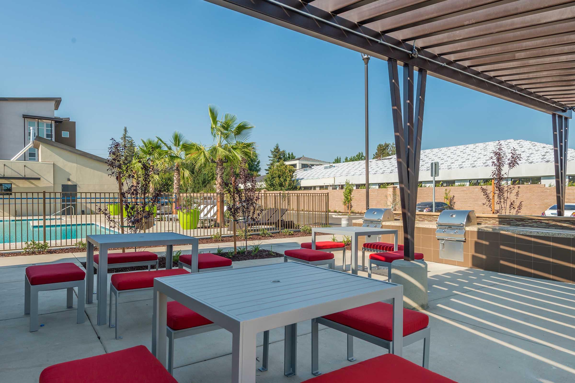 A modern outdoor seating area featuring gray metal tables with red cushions, surrounded by greenery and palm trees. In the background, there is a swimming pool and barbecue grills, with a clear blue sky overhead and buildings in the distance.