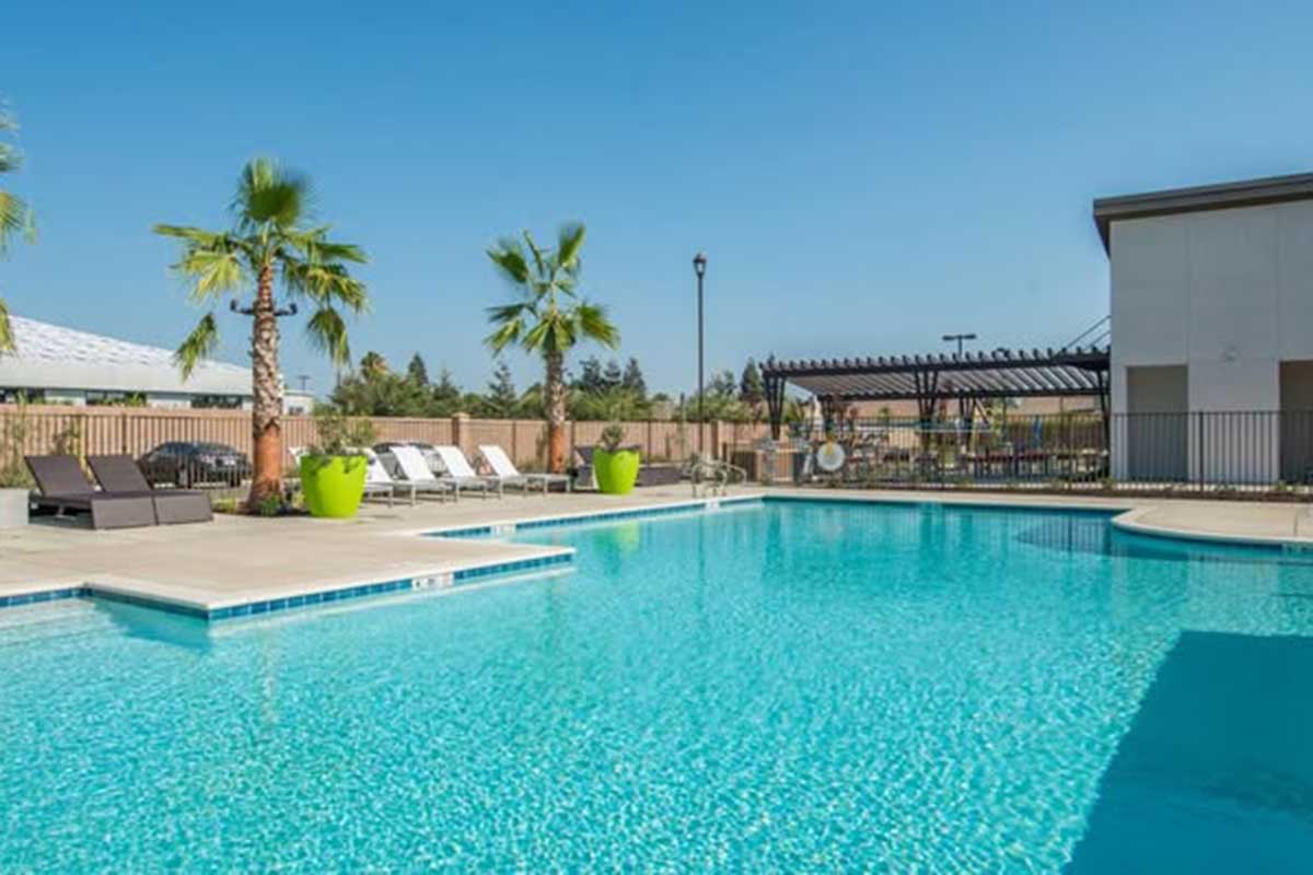 A spacious swimming pool surrounded by palm trees and lounge chairs. Bright blue water reflects the sunny sky. Green planters add a touch of color, while a shaded area is visible in the background, creating a relaxing atmosphere perfect for leisure.
