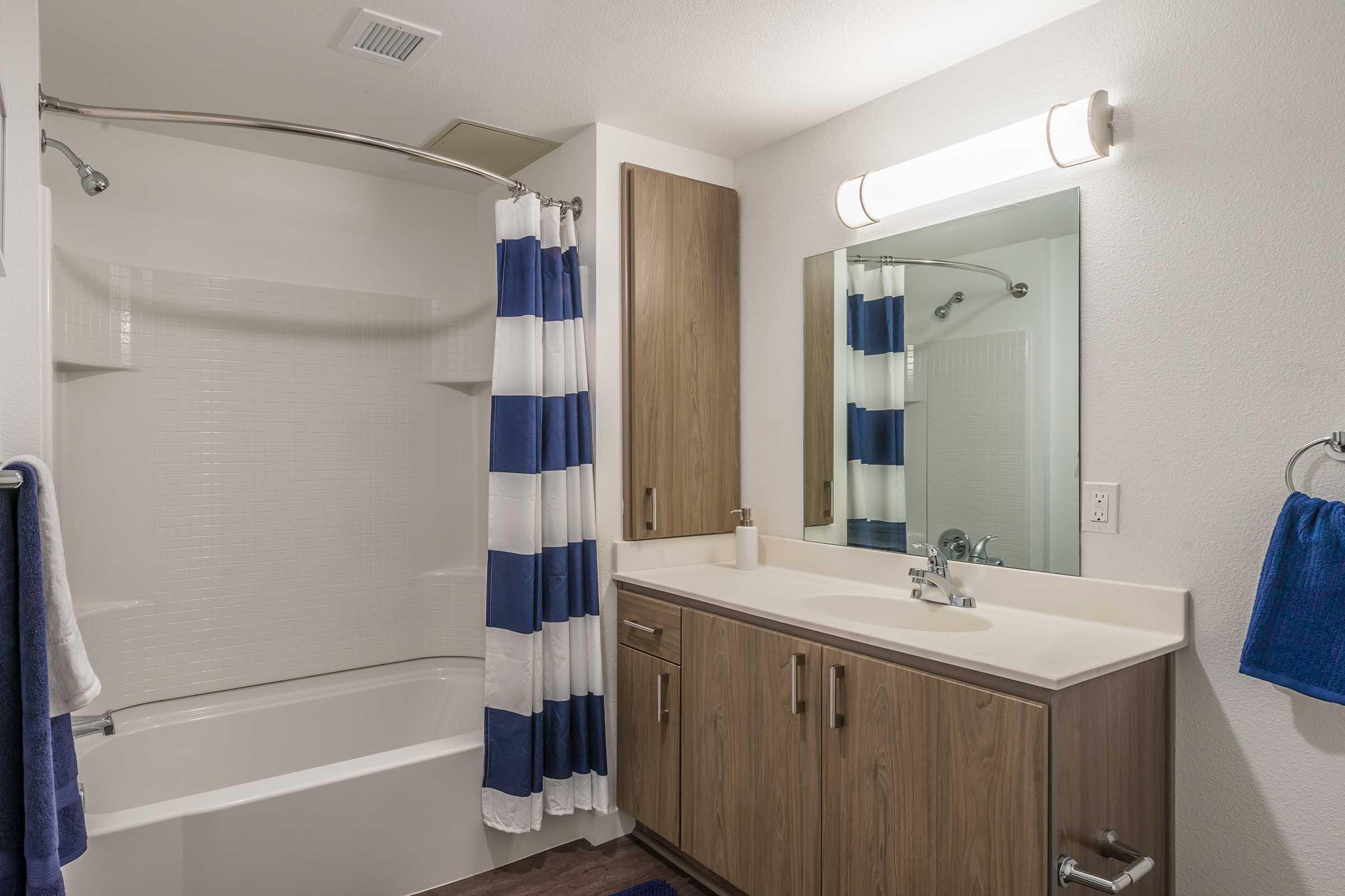 A modern bathroom featuring a white bathtub with a shower rod and striped blue and white shower curtain. The vanity has wooden cabinets with a sink and a mirror above it. A blue towel hangs on the wall, and there's a light fixture above the mirror, illuminating the space.