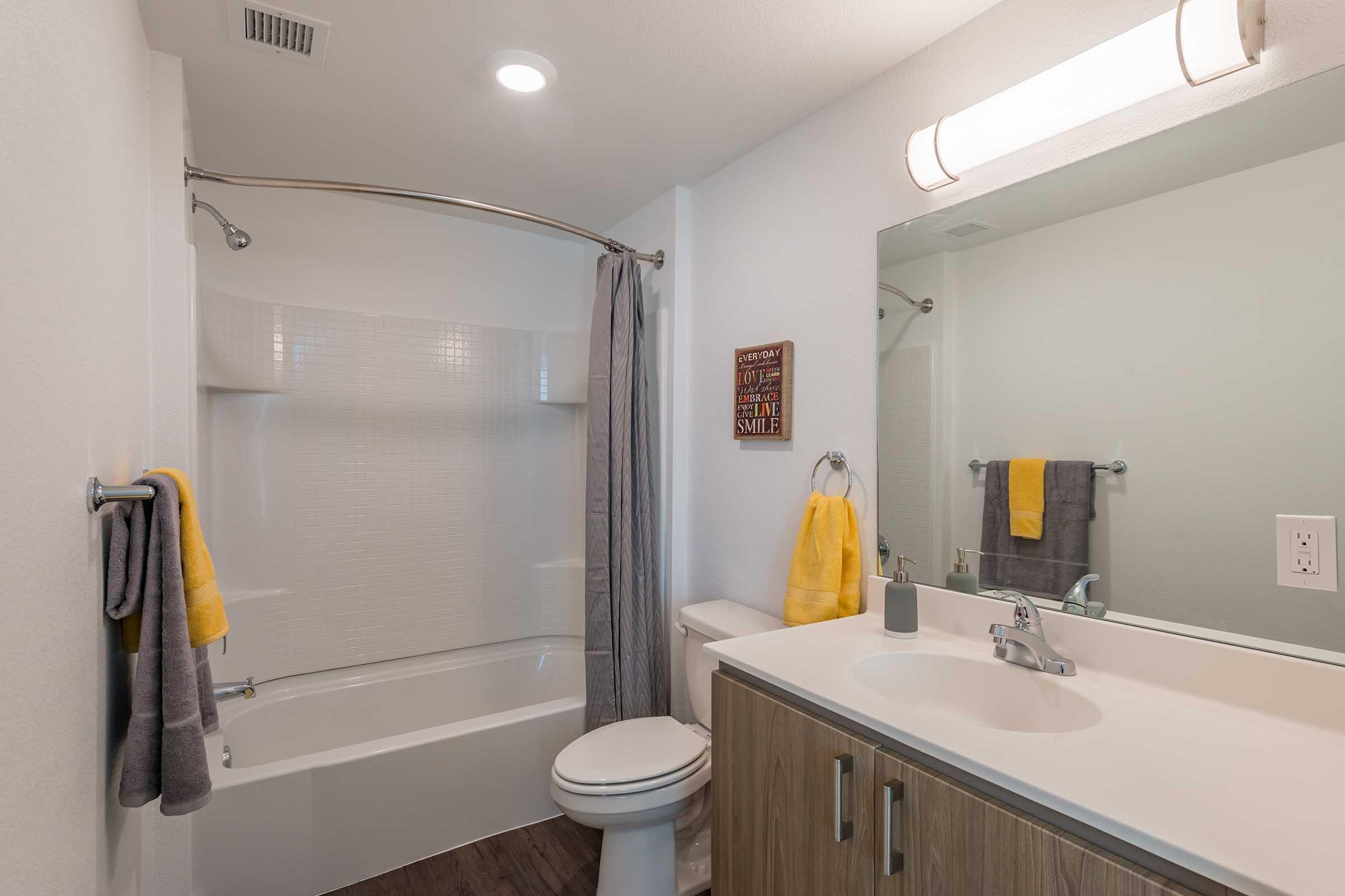 A modern bathroom featuring a bathtub with a shower curtain, a toilet, and a vanity sink. The walls are painted white, and there is a large mirror above the sink. Yellow and gray towels are hanging on towel bars. Soft lighting illuminates the space, creating a clean and inviting atmosphere.