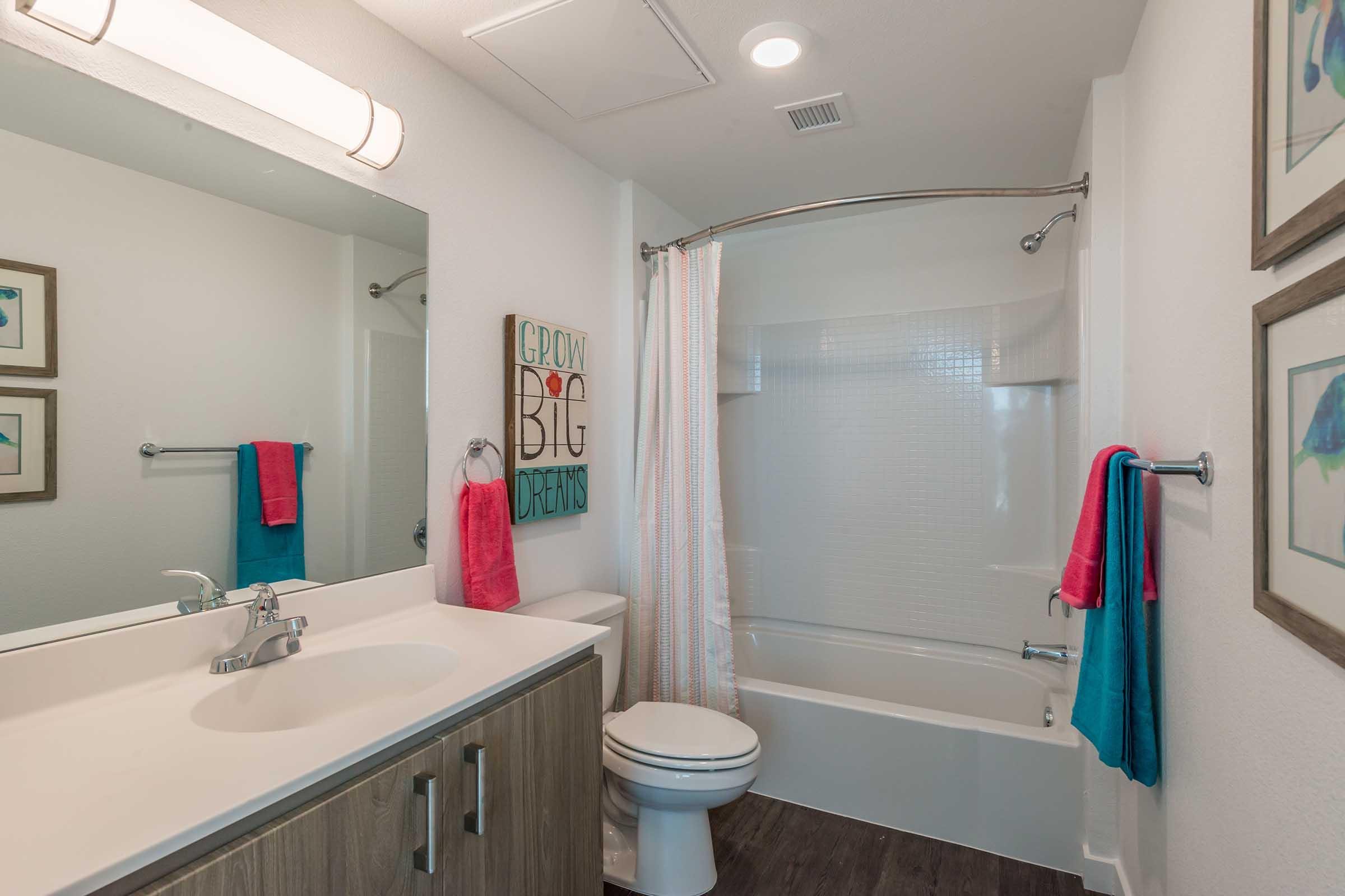 Bright and modern bathroom featuring a white bathtub with a shower curtain, a sink with a wooden vanity, and a large mirror. Colorful towels hang on a towel rack, and a wall art piece with the phrase "Grow Big Dreams" adds a whimsical touch. The overall decor is clean and inviting.