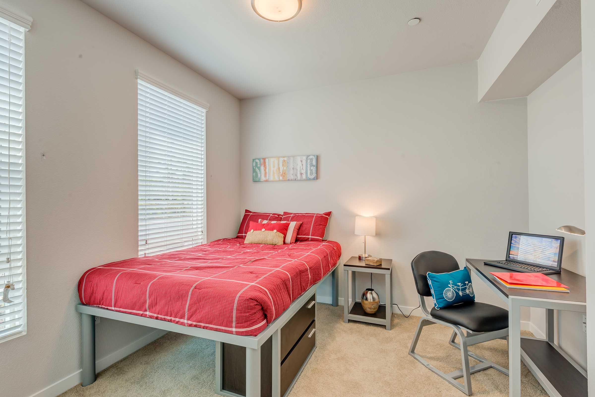Cozy bedroom with a single bed featuring red bedding, a nightstand with a lamp, and a small desk with a laptop. Natural light streams in from two windows, with light-colored walls and carpet creating a warm atmosphere. A wall decoration reads "Surfing."