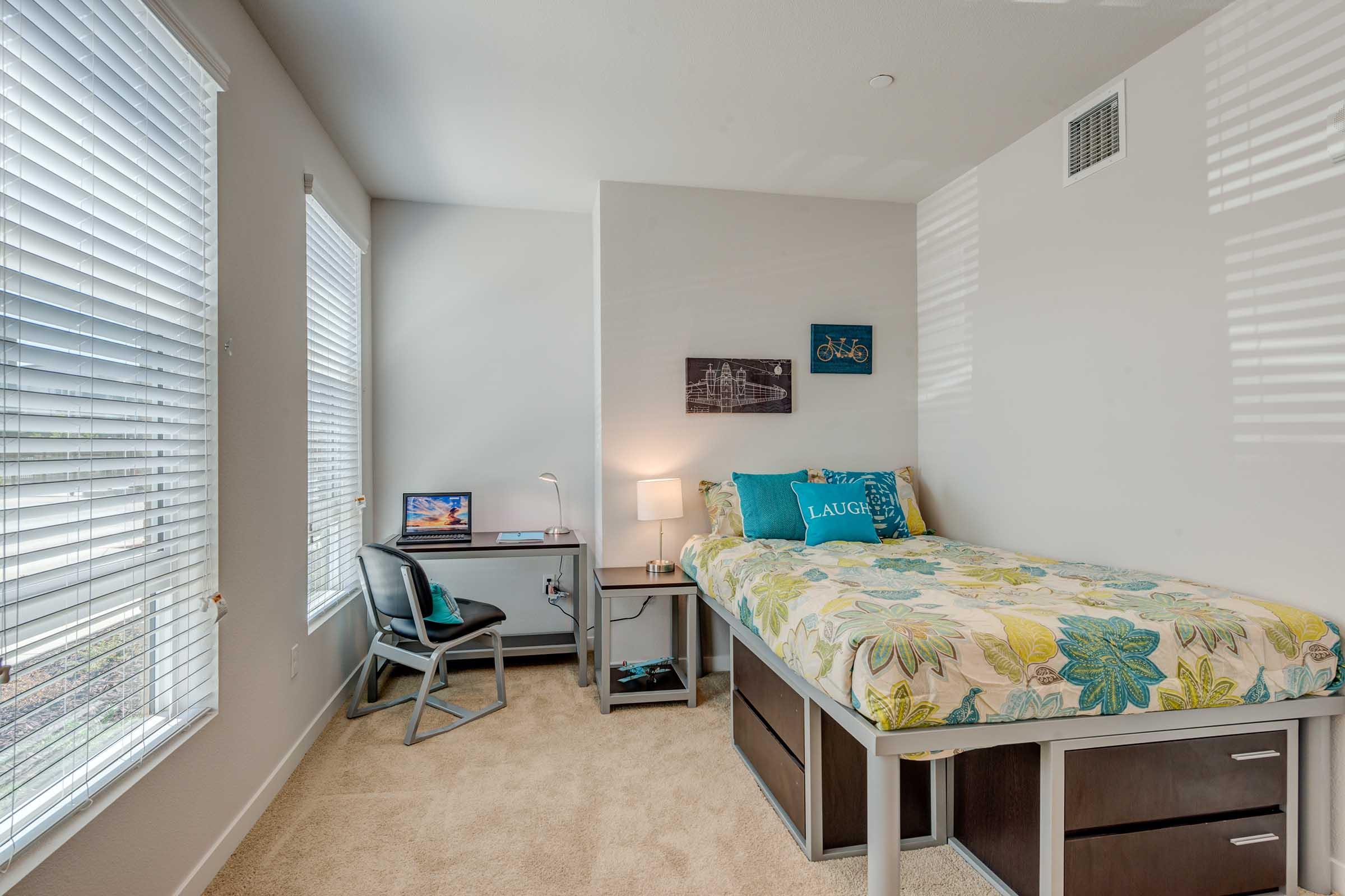 A small, well-lit bedroom featuring a single bed with floral bedding, a desk with a chair, and a lamp. There are two windows with blinds letting in natural light, and a couple of wall decorations, including a bike-themed piece and a cityscape. The room has neutral-colored walls and carpet flooring.