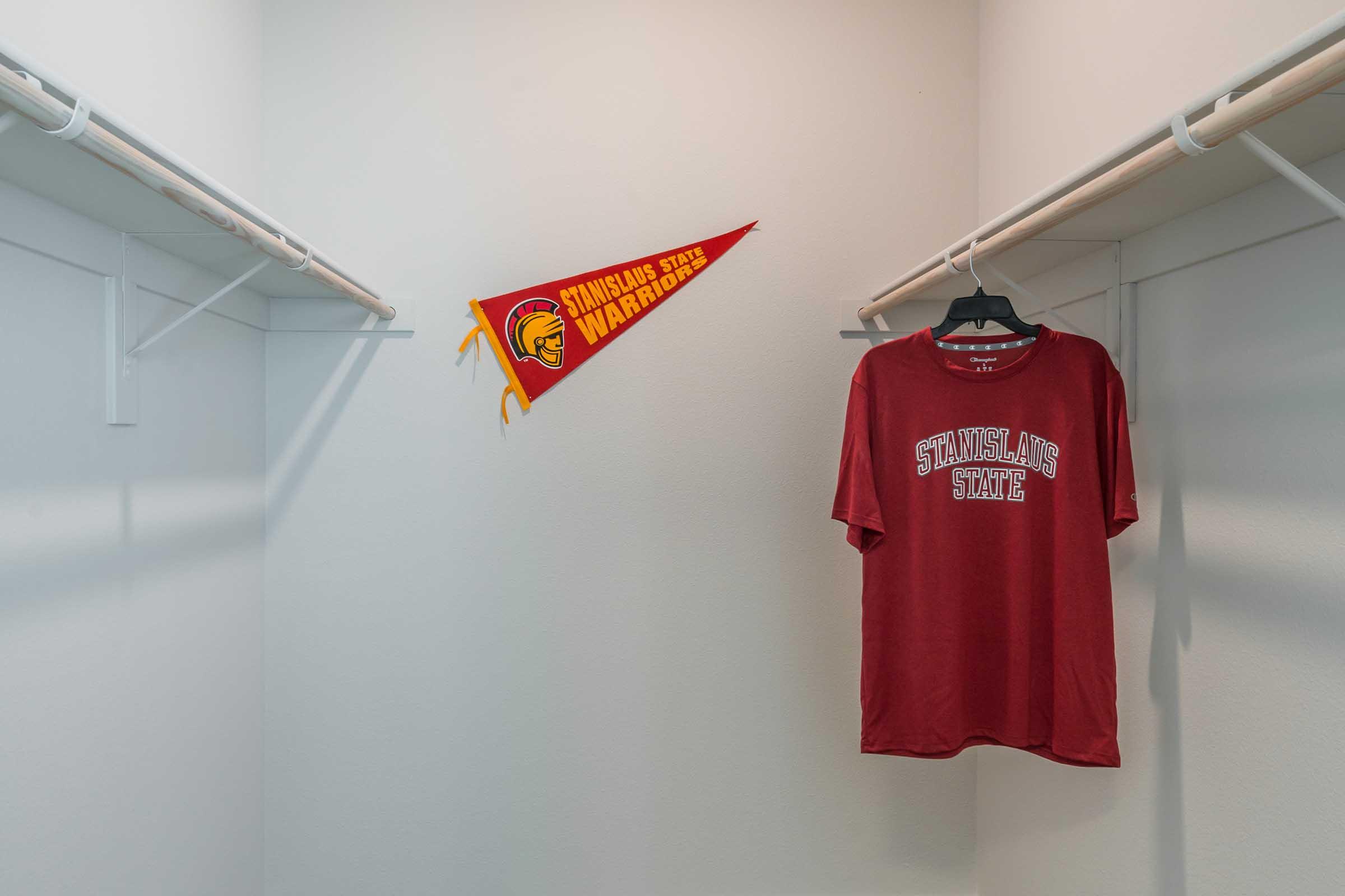 A red Stanislaus State Warriors shirt hanging on a black hanger inside a closet, with a red pennant showcasing the Stanislaus State logo and the team name attached to the wall. The closet has white walls and a minimalistic design.