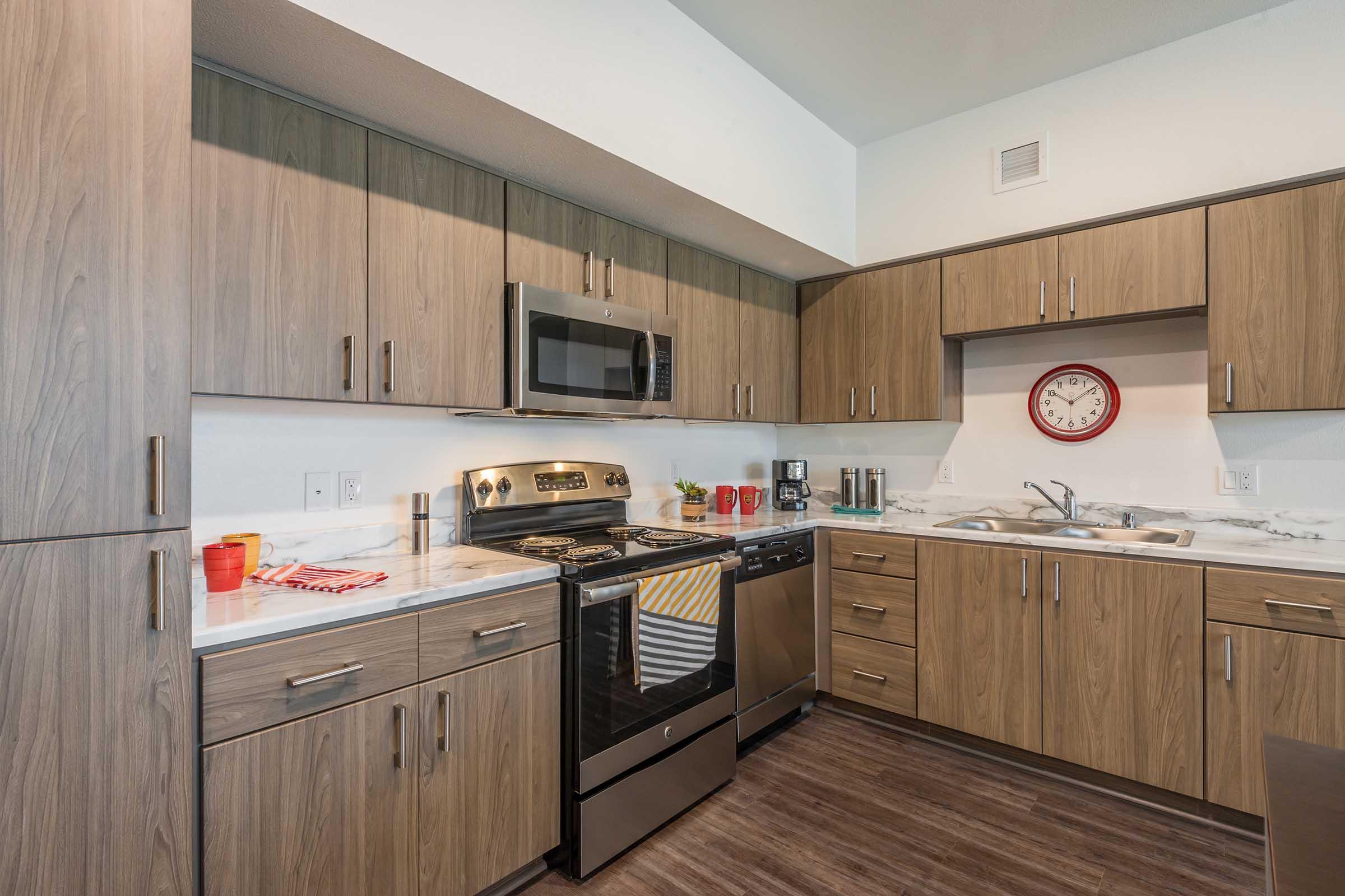 Modern kitchen featuring wooden cabinetry, stainless steel appliances, and a marble countertop. The layout includes a stove, microwave, and sink, with a wall clock and small kitchen accessories. The flooring is a dark wood, creating a warm and inviting atmosphere.