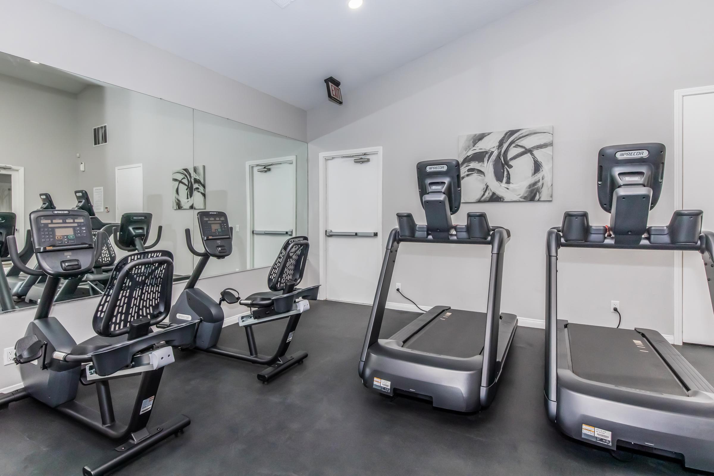 A modern gym interior featuring exercise equipment, including recumbent bikes and treadmills, on a black floor. The walls are painted light grey, and a large mirror reflects the equipment. Abstract wall art is visible in the background, enhancing the contemporary atmosphere of the fitness space.