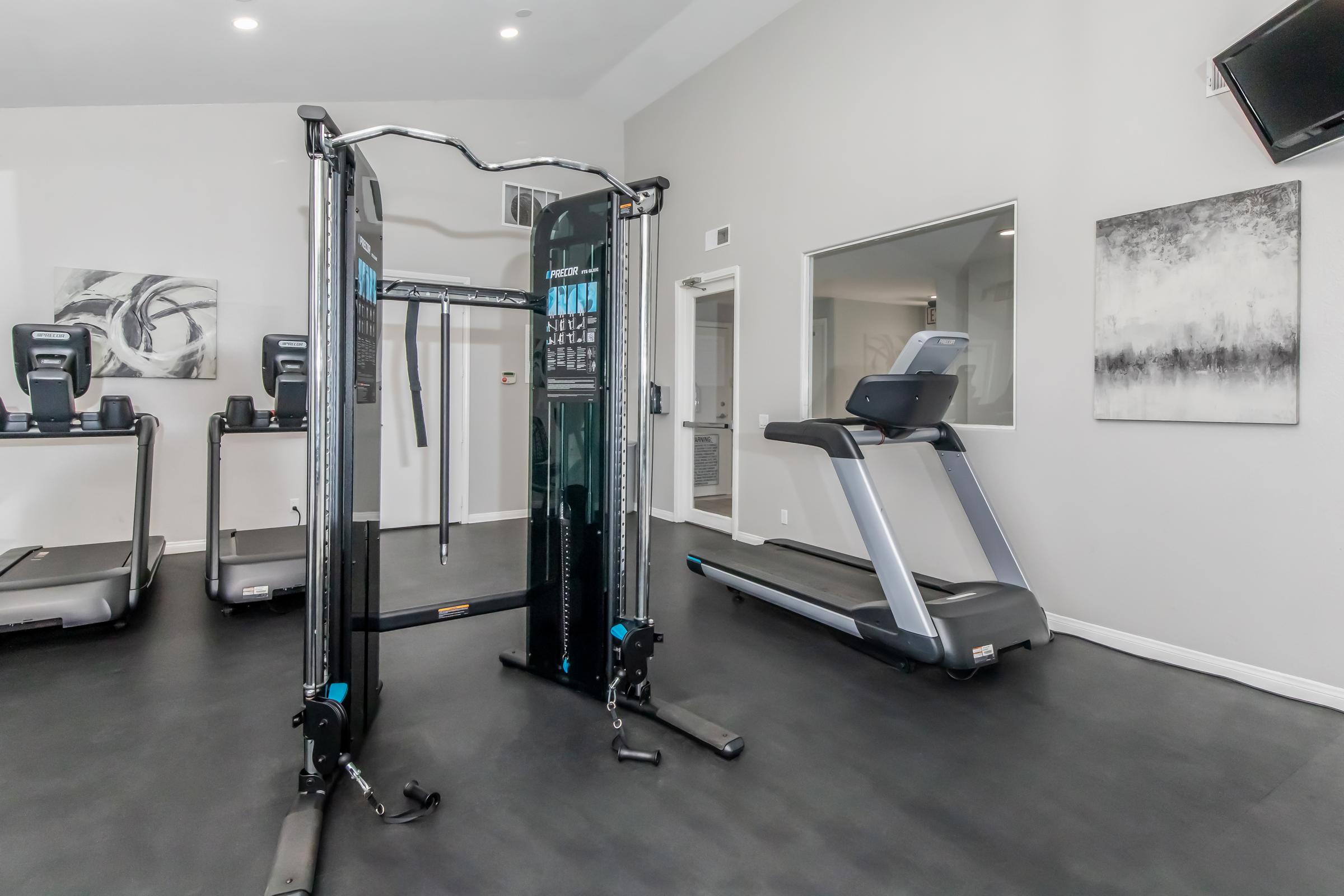 A modern gym interior featuring a cable machine and a treadmill. The room has light gray walls and a large window, allowing natural light to enter. There are exercise machines in the background, with minimalist artwork on the walls and overhead lighting enhancing the space's bright and inviting atmosphere.