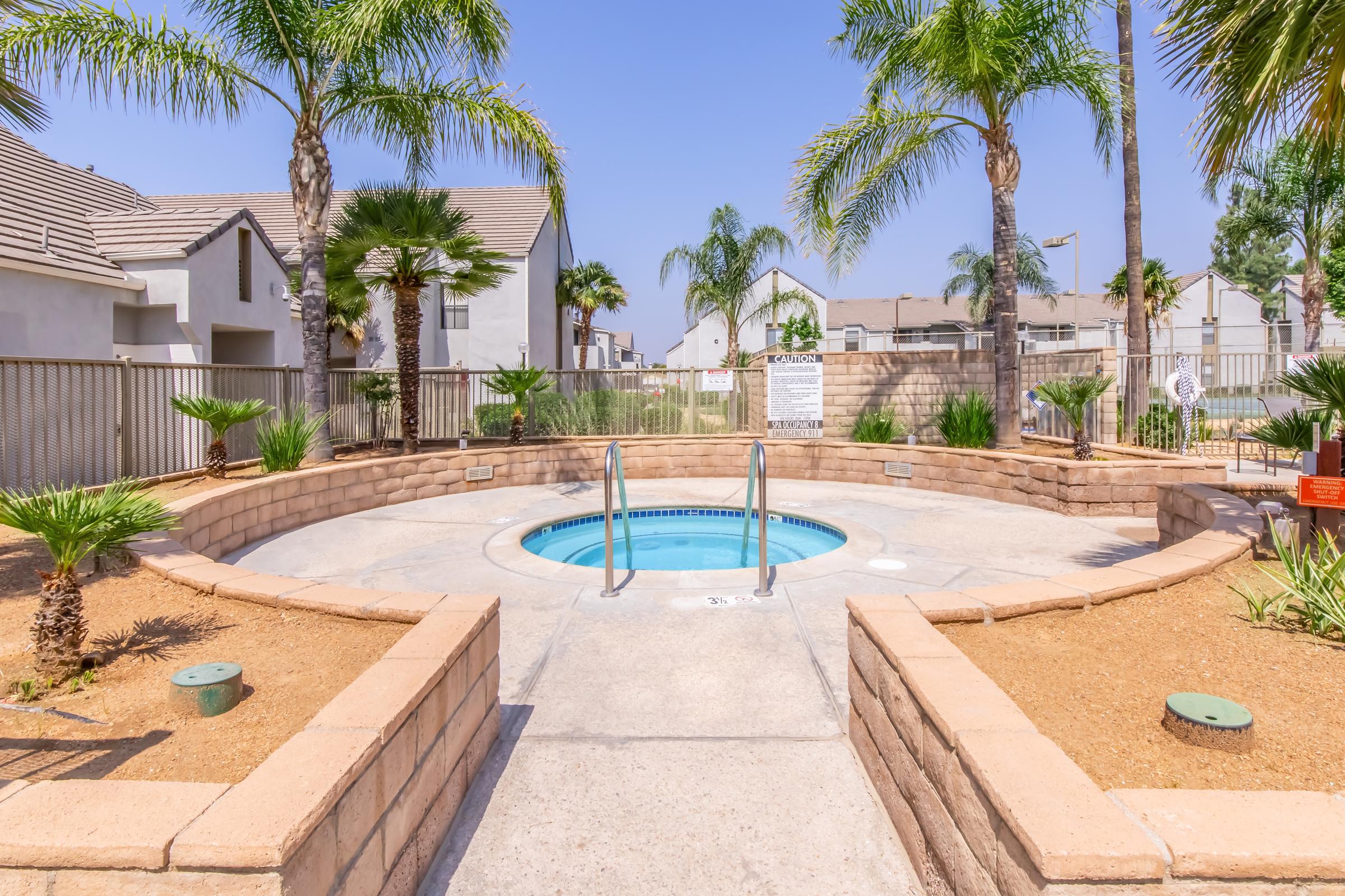 A small, circular hot tub surrounded by palm trees and dry landscaping. The area is enclosed by a low fence, with residential buildings in the background under a clear blue sky. There are steps leading into the hot tub and plants evenly spaced around the concrete surface.