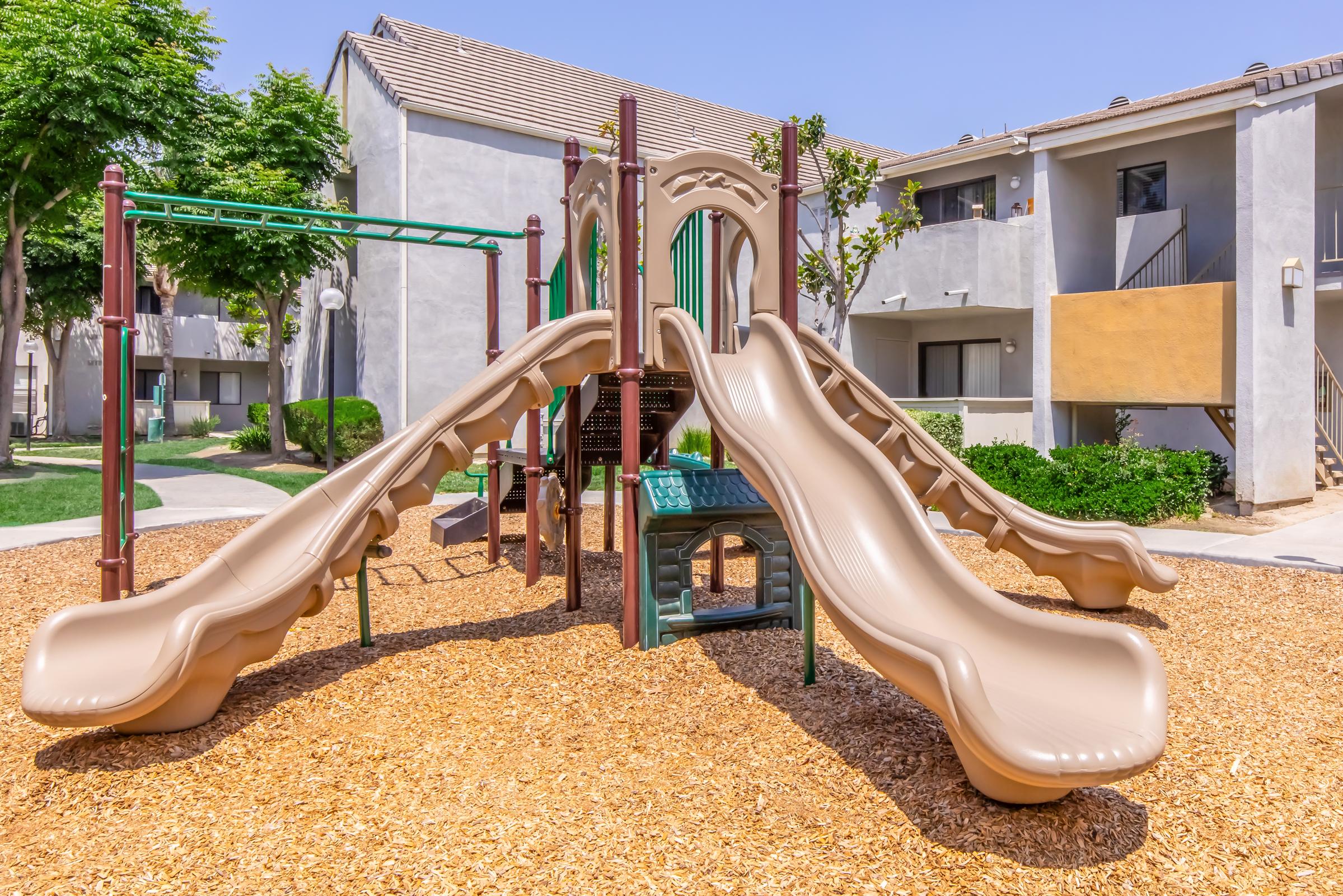Playground structure featuring two large brown slides, a climbing frame, and interactive panels. Surrounding the playground are green trees and residential buildings. The ground is covered with wood mulch, creating a soft play area. The setting is bright and sunny, indicating a pleasant day.