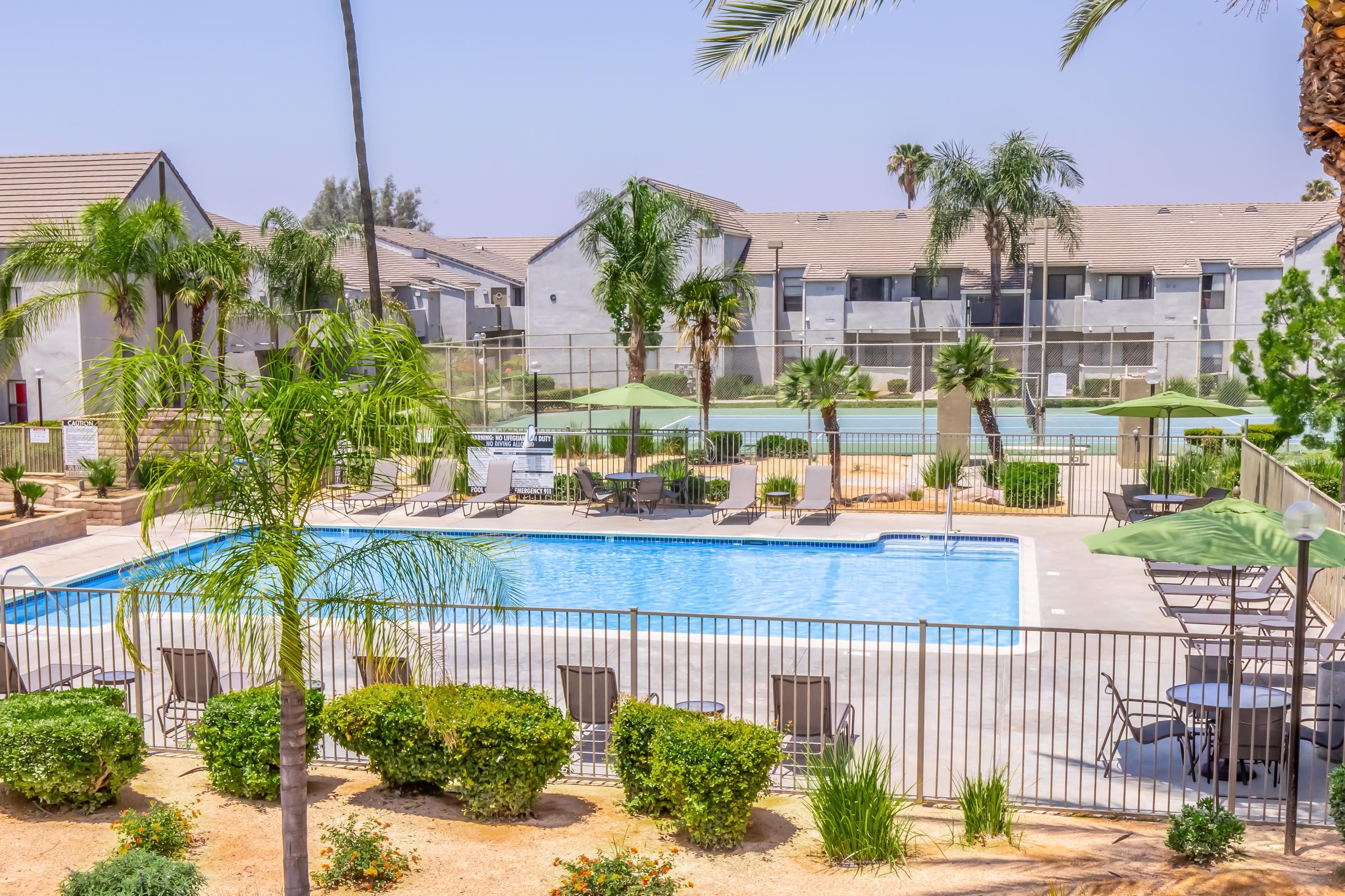 A brightly lit swimming pool surrounded by palm trees and lounge chairs. There are green umbrellas for shade, and in the background, there are multi-story residential buildings and a tennis court. The scene is serene and inviting, ideal for relaxation and leisure activities.