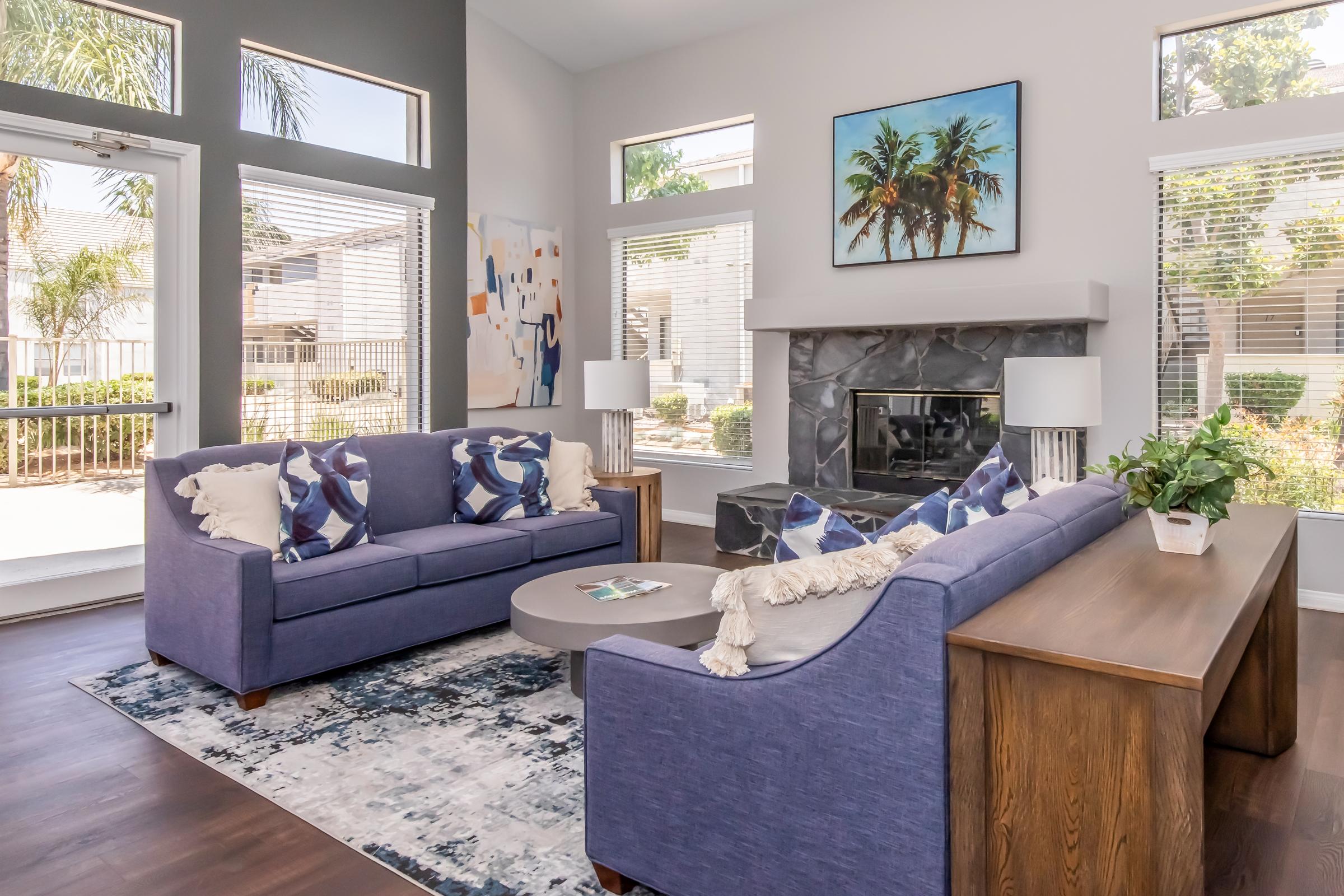 A bright and modern living room featuring two blue sofas with decorative pillows, a wooden coffee table, and a fireplace. Large windows allow natural light to fill the space, which is adorned with plants and art on the walls. A cozy and inviting atmosphere is created with stylish decor and a soft area rug.