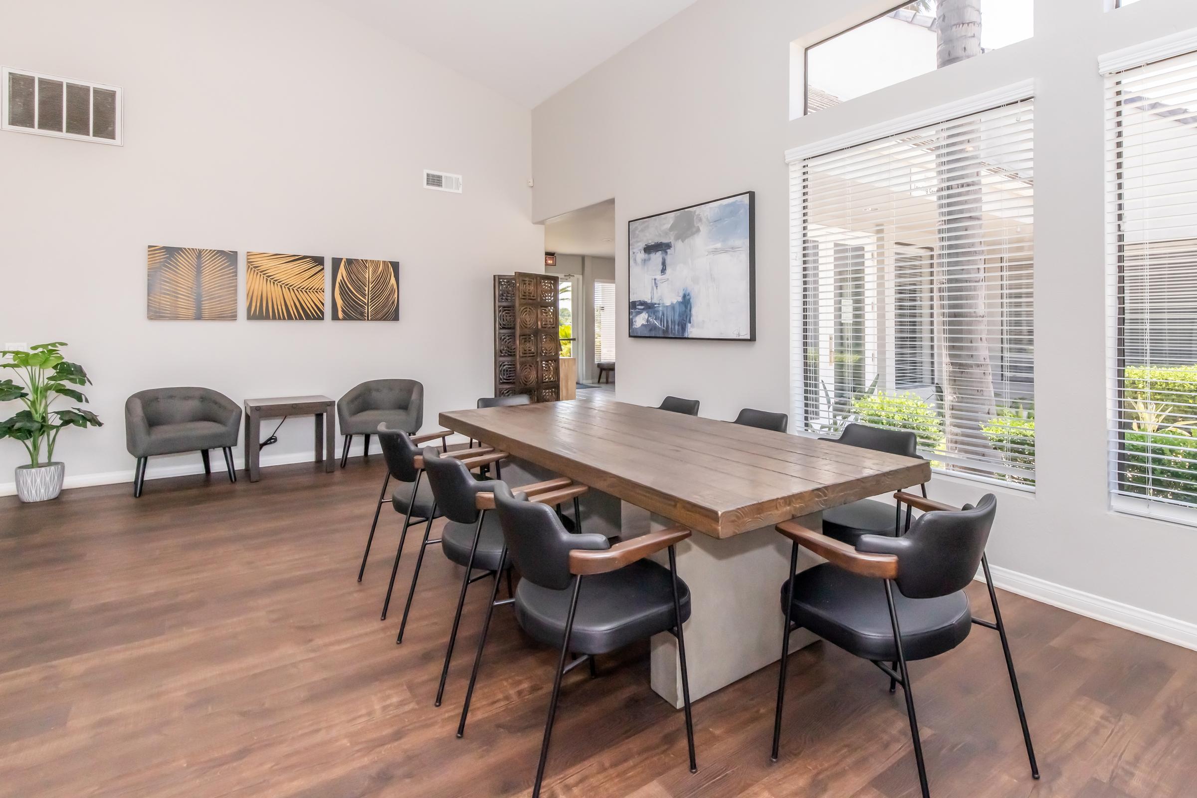 A modern meeting room featuring a spacious wooden table with concrete legs surrounded by black chairs. The room has large windows letting in natural light, and decor includes three framed artworks on the wall and a potted plant in the corner. The floor is made of dark wood.