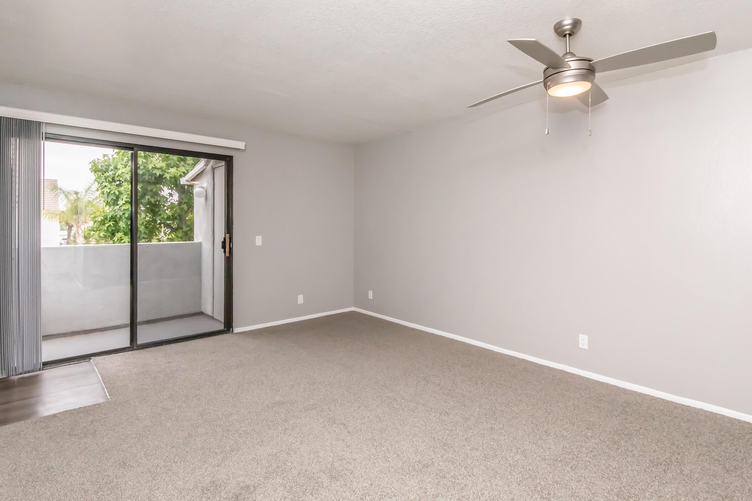 Spacious, empty living room featuring light gray walls and carpet. A ceiling fan hangs in the center, and there is a large sliding glass door leading to a balcony. Natural light illuminates the room from outside.