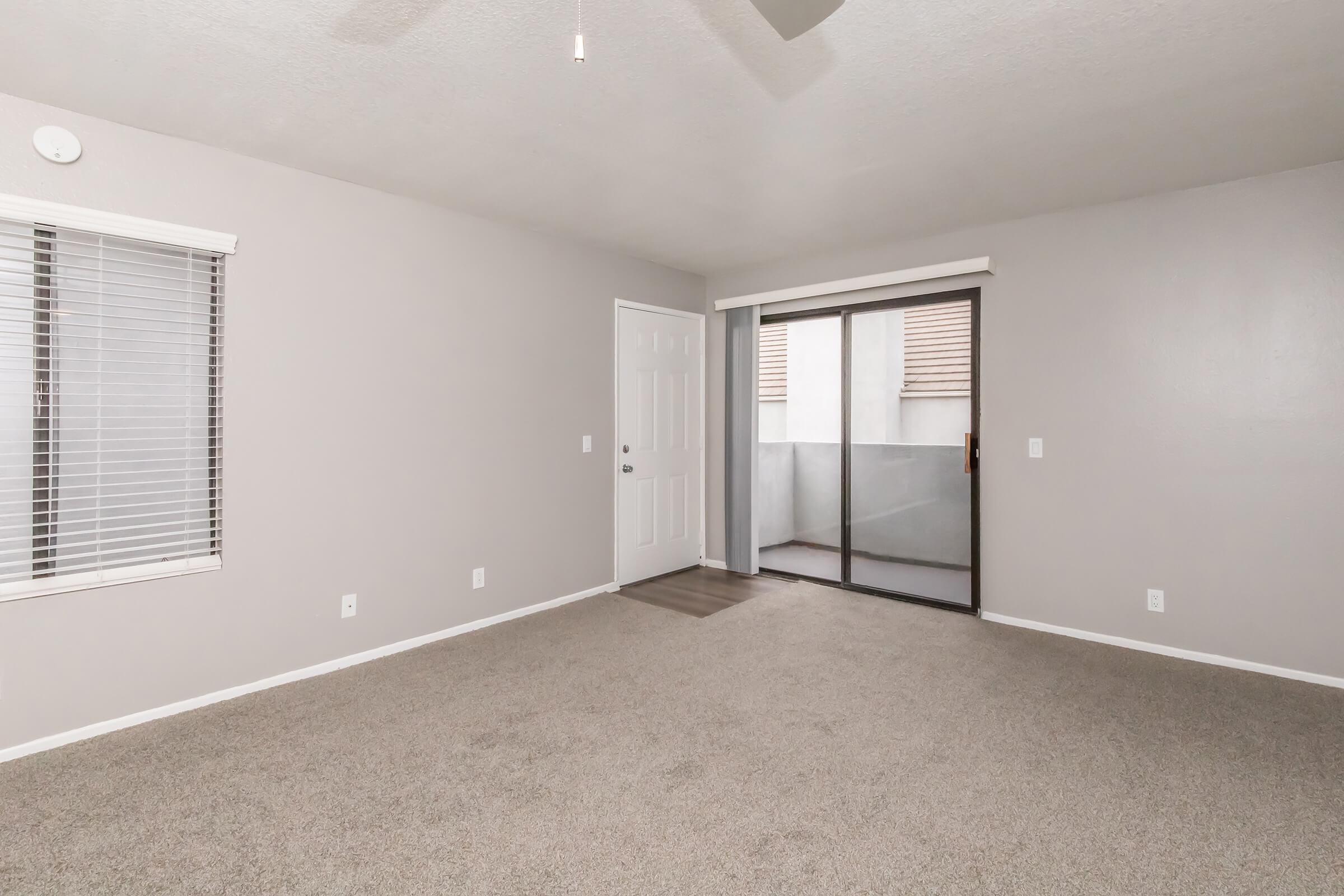 A spacious, vacant living room with light gray walls and a plush carpet. There is a ceiling fan, a closed door leading outside, and large windows with blinds that allow natural light to enter. The room features minimal decor, creating a bright and airy atmosphere.
