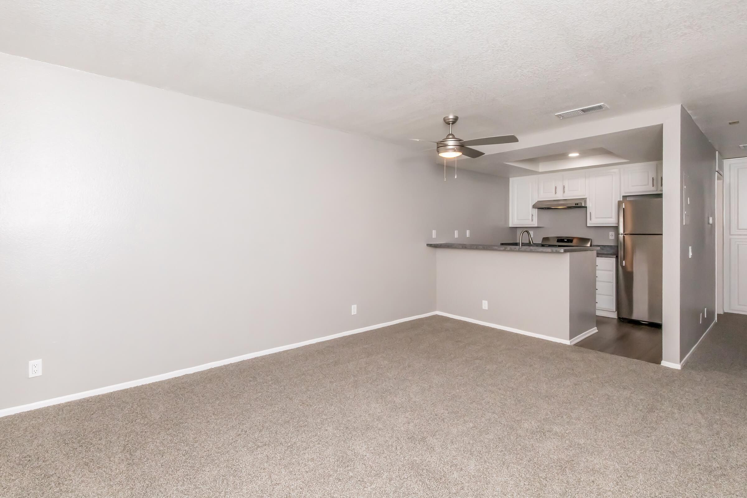 Spacious, carpeted living area with light gray walls and a ceiling fan. Adjacent to a modern kitchen featuring white cabinetry, stainless steel appliances, and a breakfast bar. The layout creates an open and inviting atmosphere.