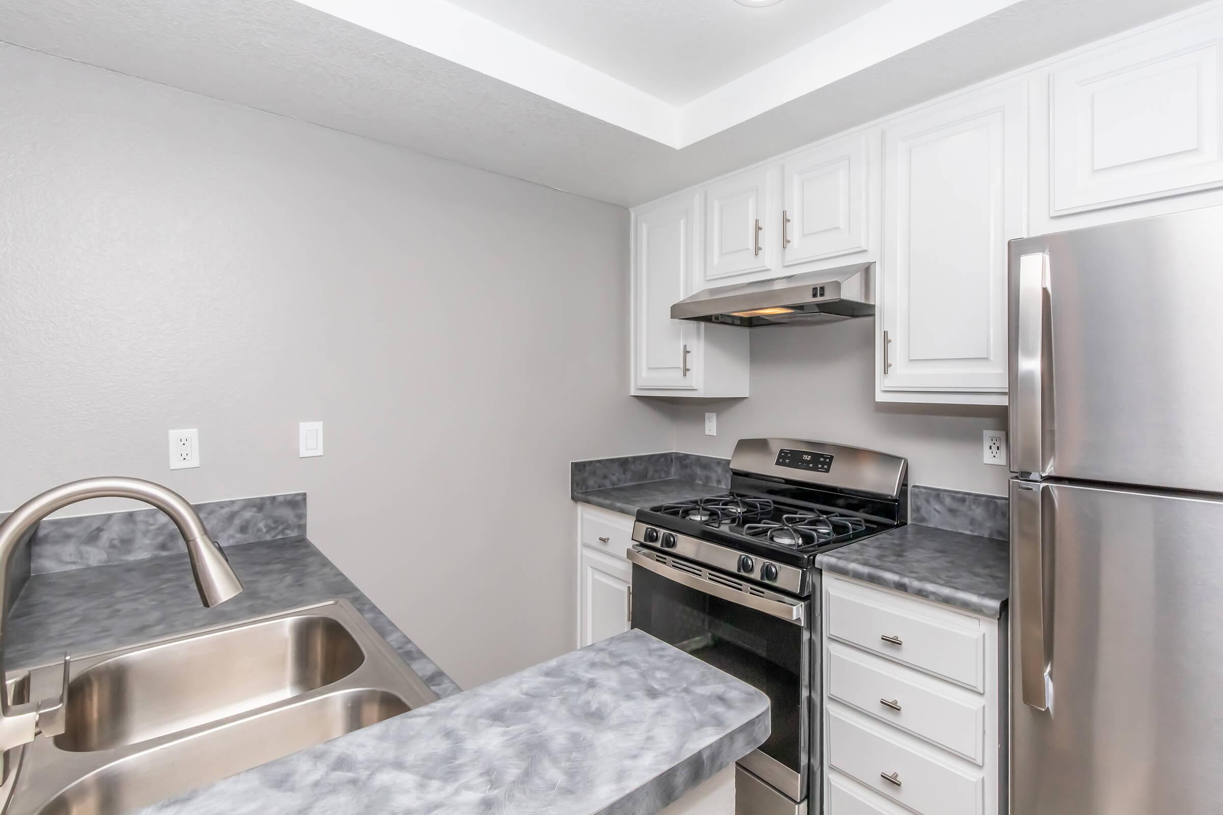 Modern kitchen featuring white cabinets, gray countertops, a stainless steel gas stove, an overhead range hood, and a stainless steel refrigerator. The space is well-lit with a neutral gray wall color and includes a double sink with a sleek faucet.
