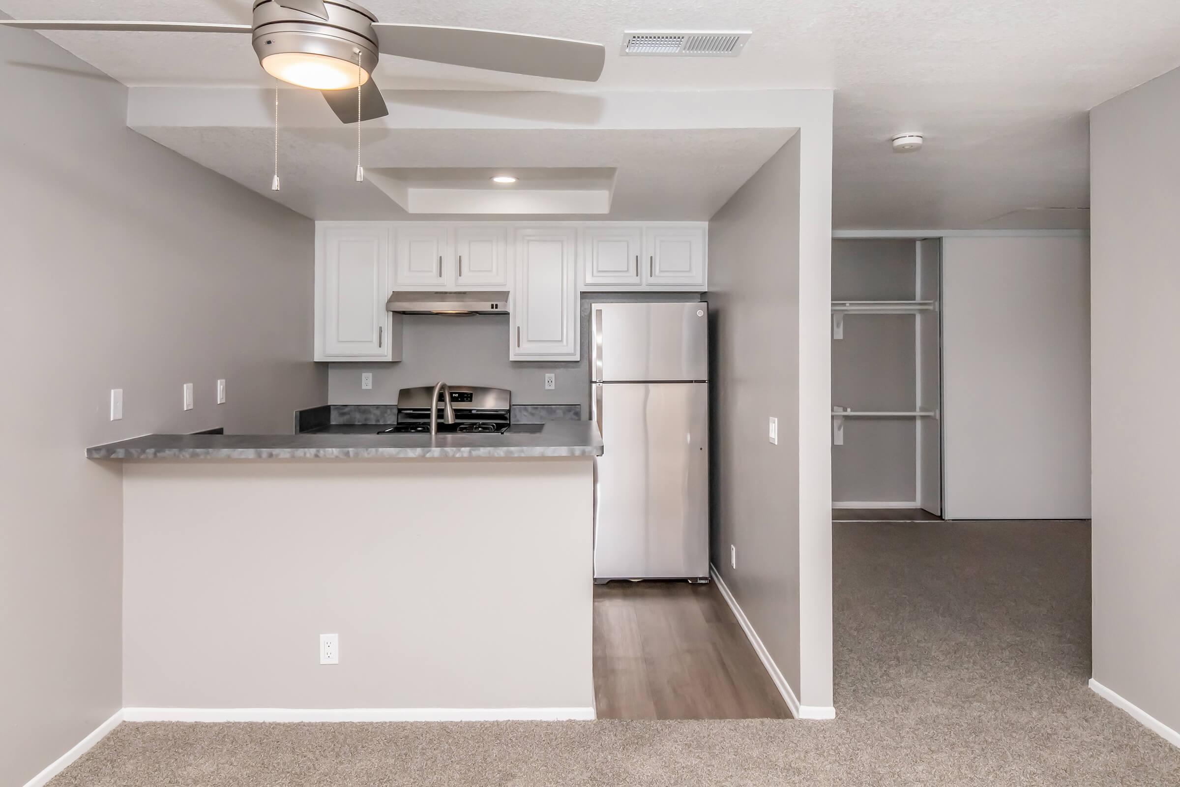 A modern kitchen with white cabinets and a stainless steel refrigerator, featuring a gray countertop and an open layout. There's a ceiling fan above and a view into a spacious room with carpeted flooring and an empty closet on the right. The walls are painted in a light gray color.