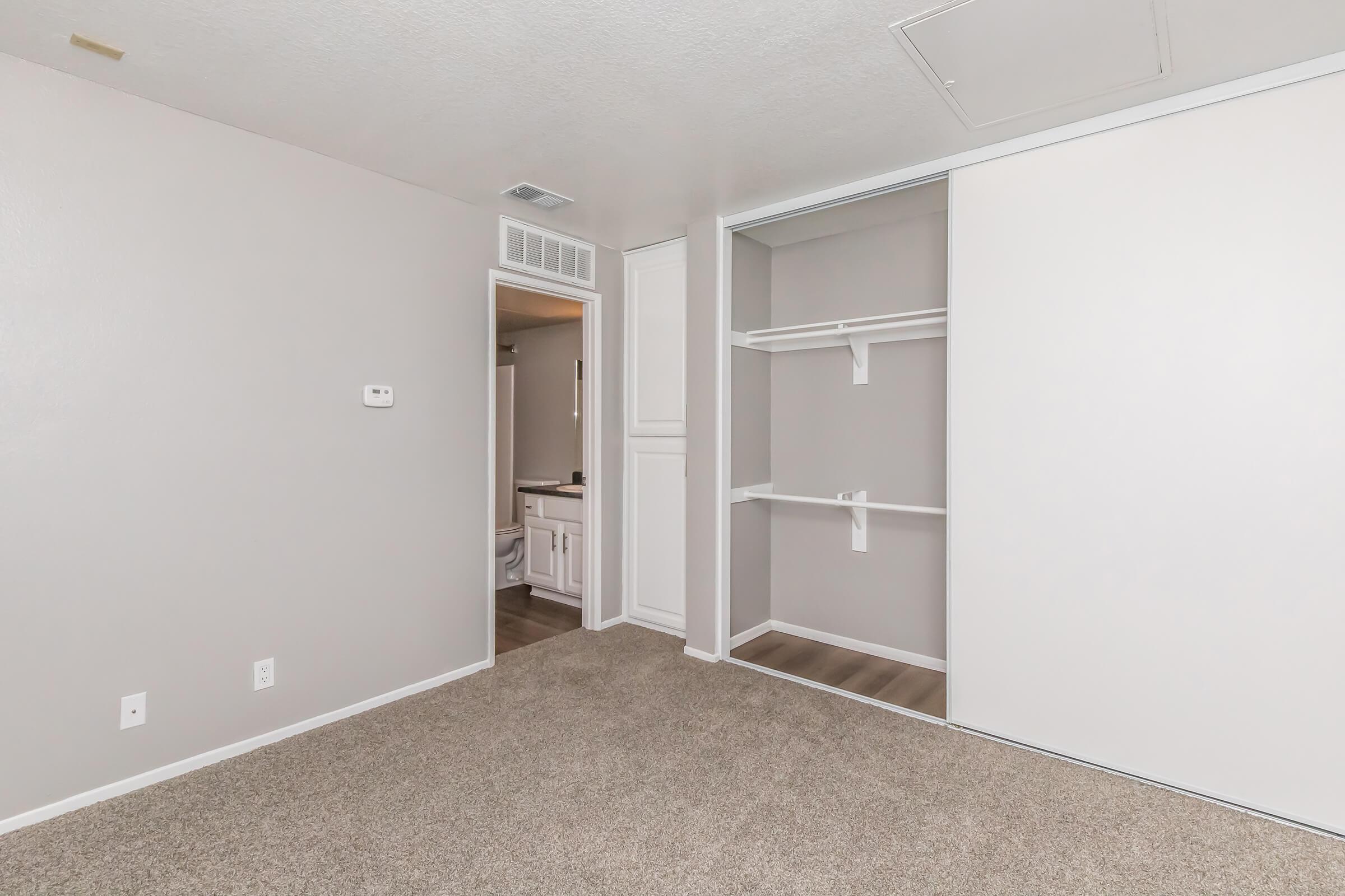 A light gray room with beige carpet, featuring a small closet with shelving. A doorway leads to a bathroom, and there is a wall-mounted thermostat. The walls are plain, and there is a ceiling vent. Overall, the space appears bright and uncluttered.