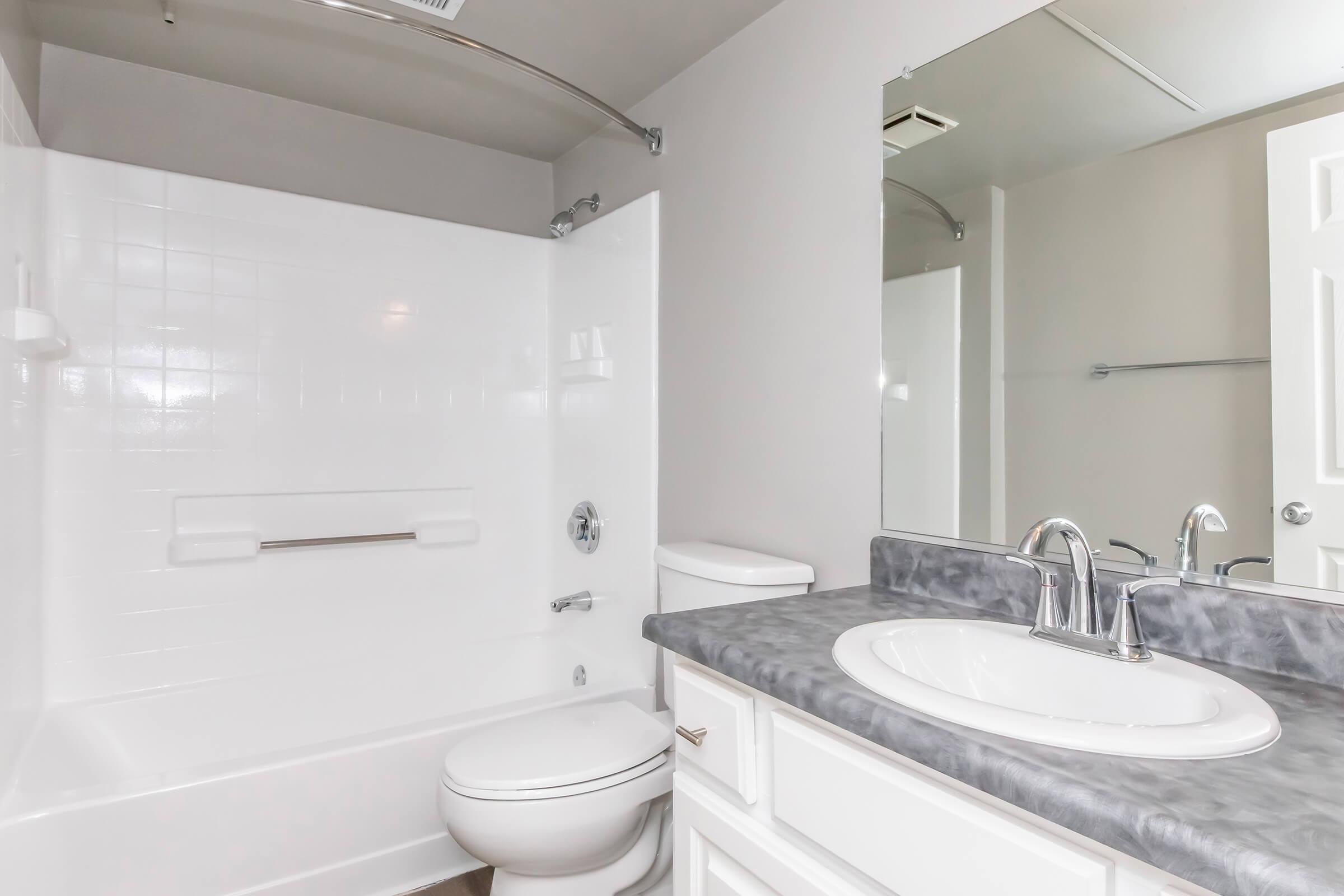 A clean, modern bathroom featuring a white tub and shower combination, a sleek sink with a gray countertop, a toilet, and a large mirror. The walls are painted in a neutral tone, and there's ambient lighting in the space.