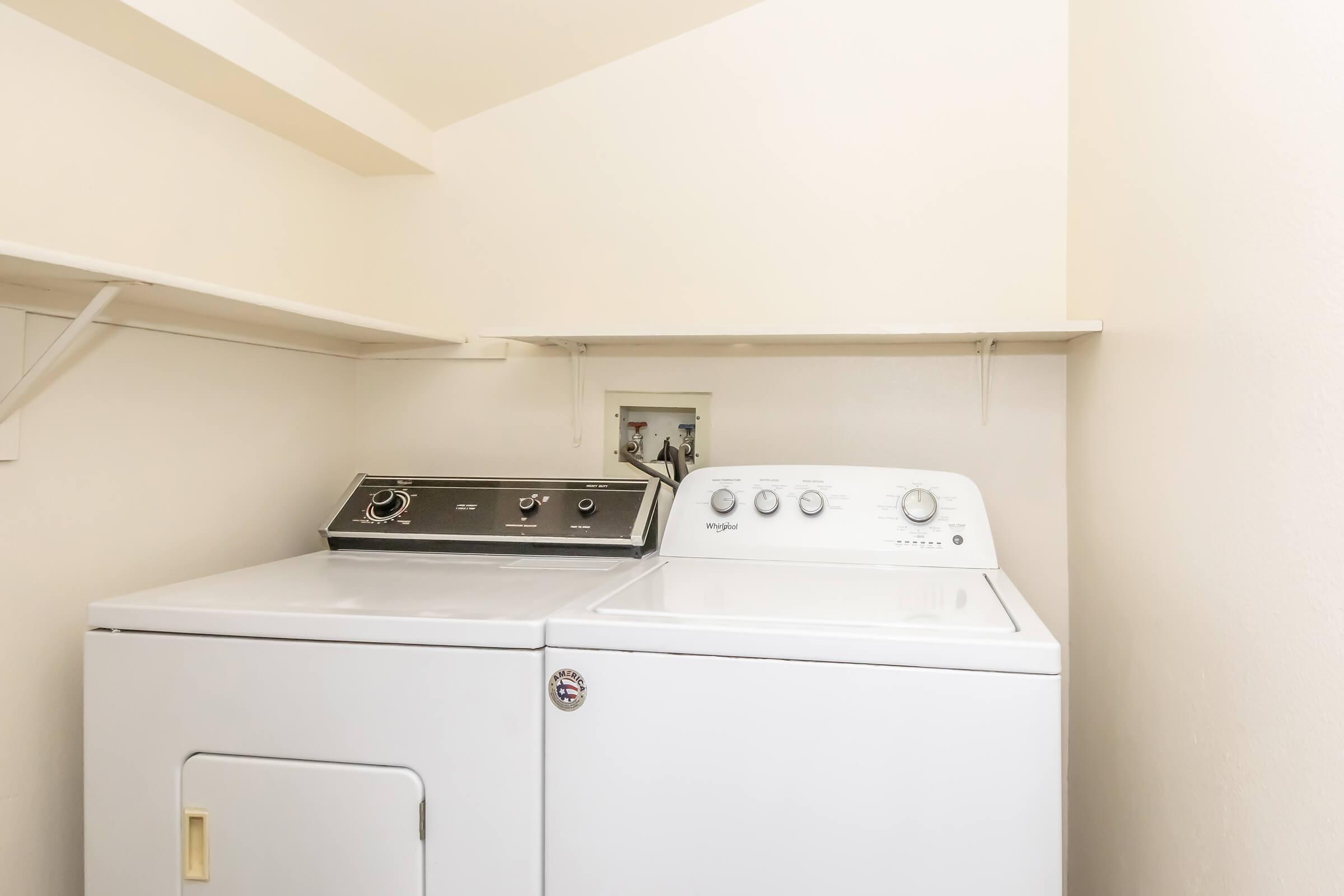 A laundry room with two washing machines, one on the left with a retro design and knobs, and a white washing machine on the right. There is a shelf mounted above the machines and a blank wall behind them, creating a clean and functional laundry space.